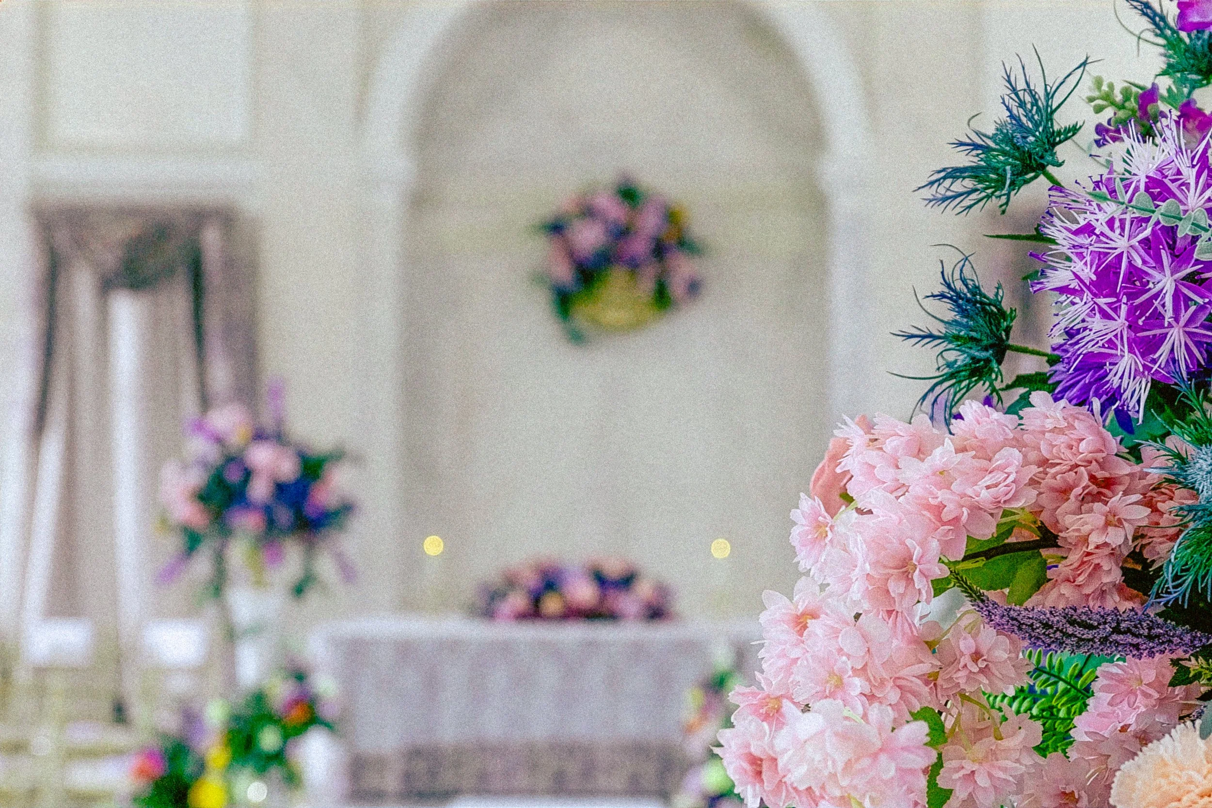 Colorful floral arrangement with pink and purple flowers in the foreground, blurred pastel-colored decorations in the background.