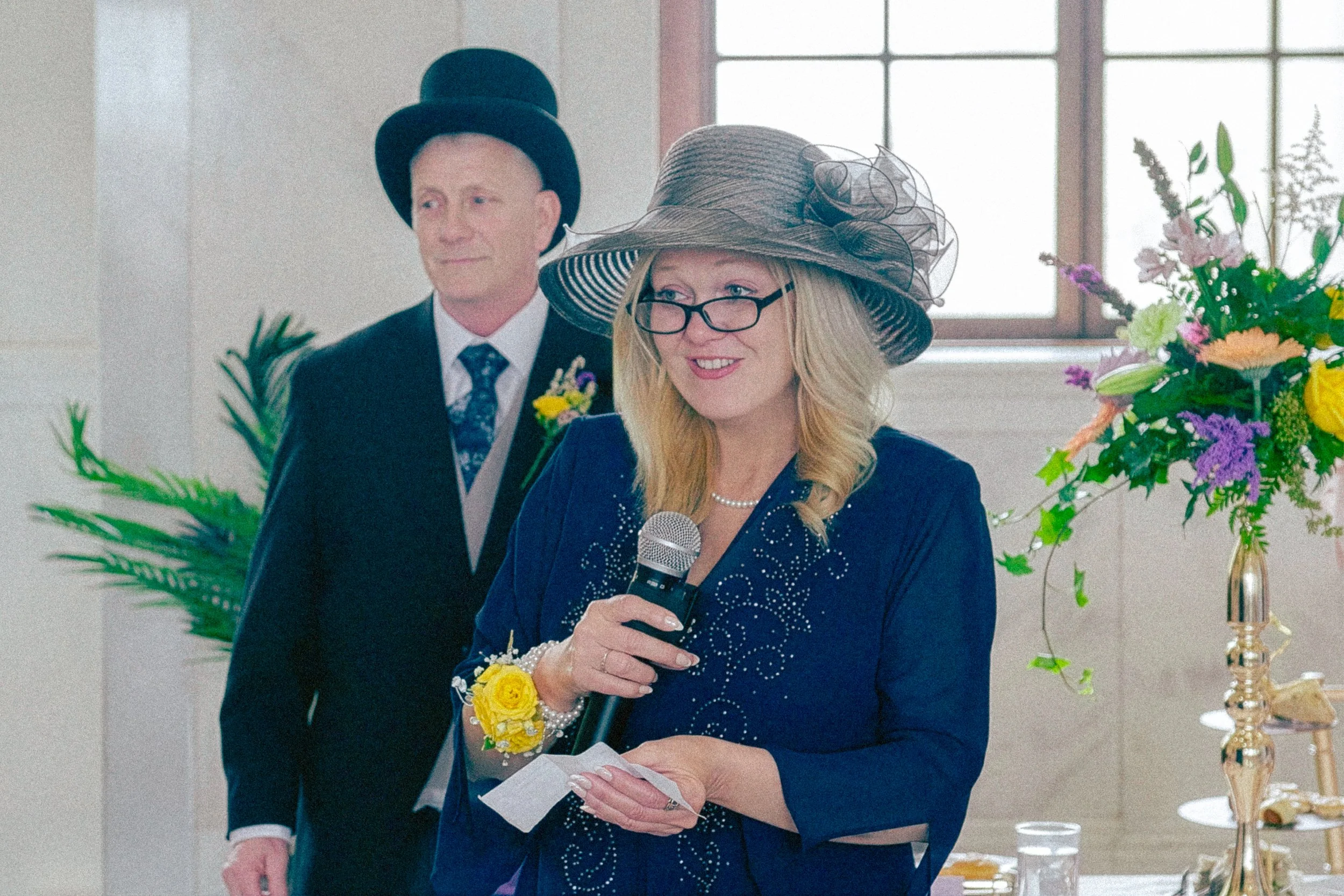A woman wearing glasses and a large decorated hat speaking into a microphone at a celebration or event, with a man in a suit and top hat standing behind her, and floral arrangements on a table nearby.