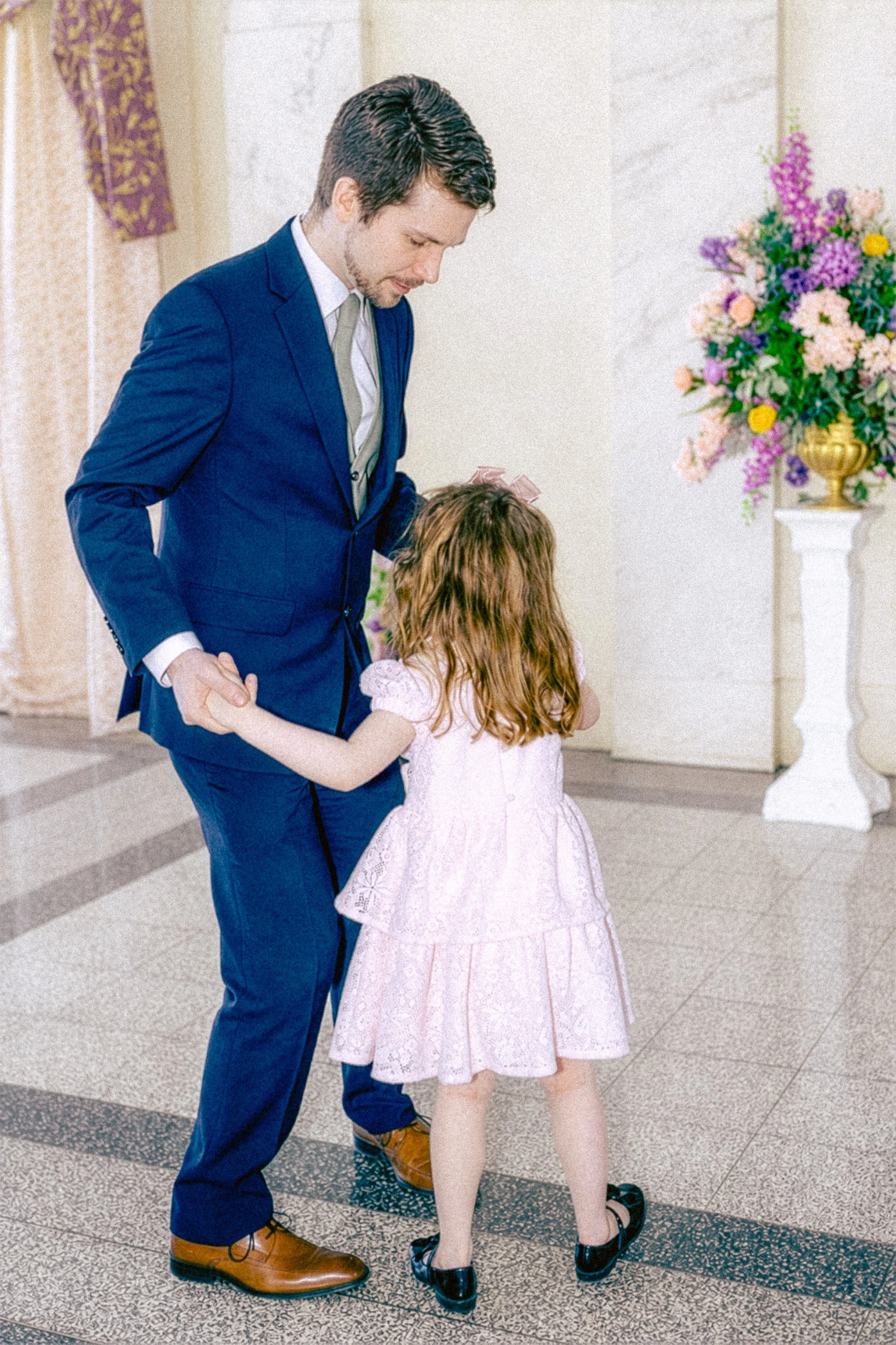 A man in a blue suit dancing with a young girl in a pink dress at an indoor event with floral arrangements nearby.
