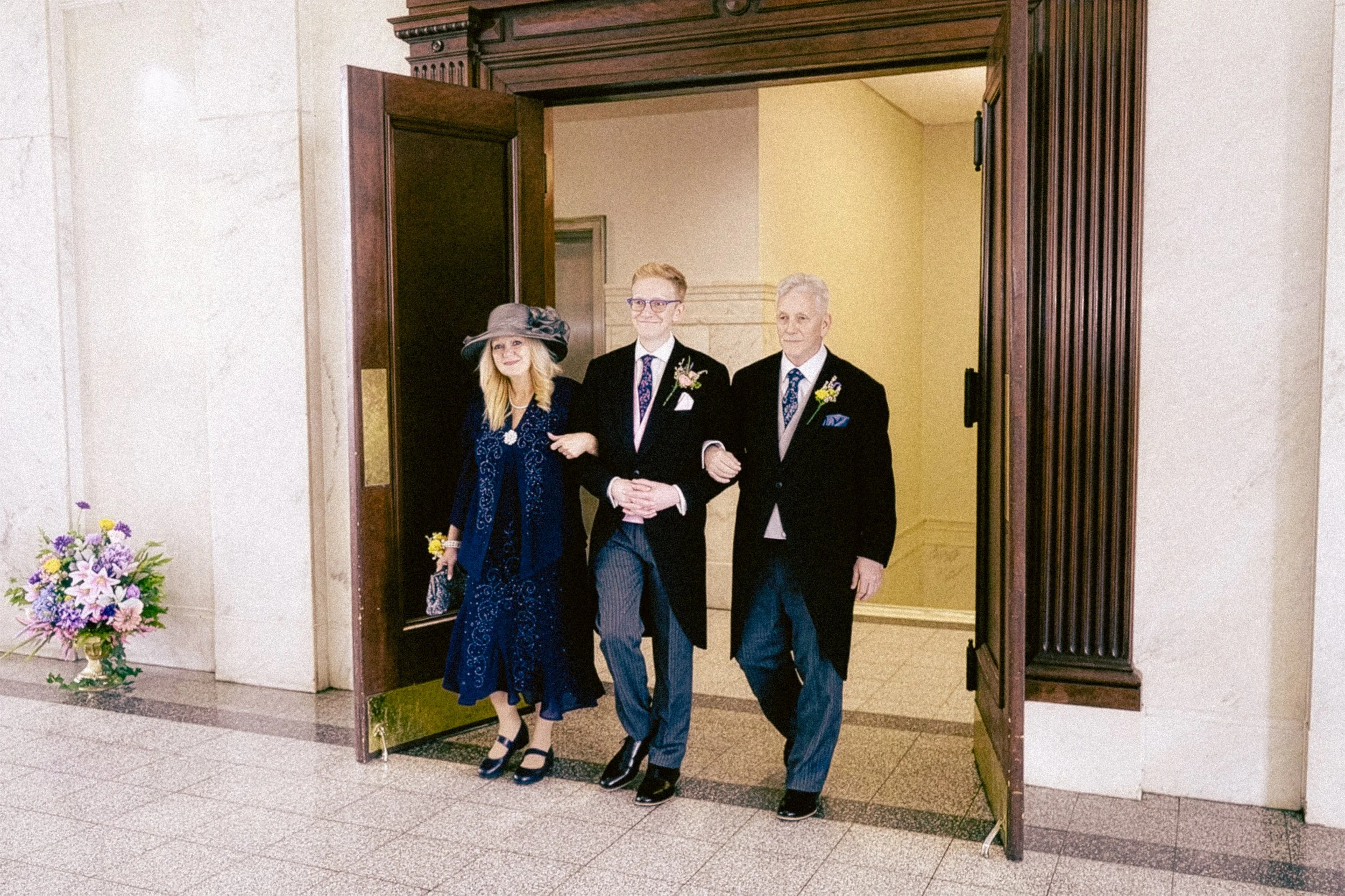 Three people, two men and a woman, walking out of a building through a wooden doorway, dressed formally, with flowers on the woman's wrist and on the men's lapels, indicating a wedding or special event.