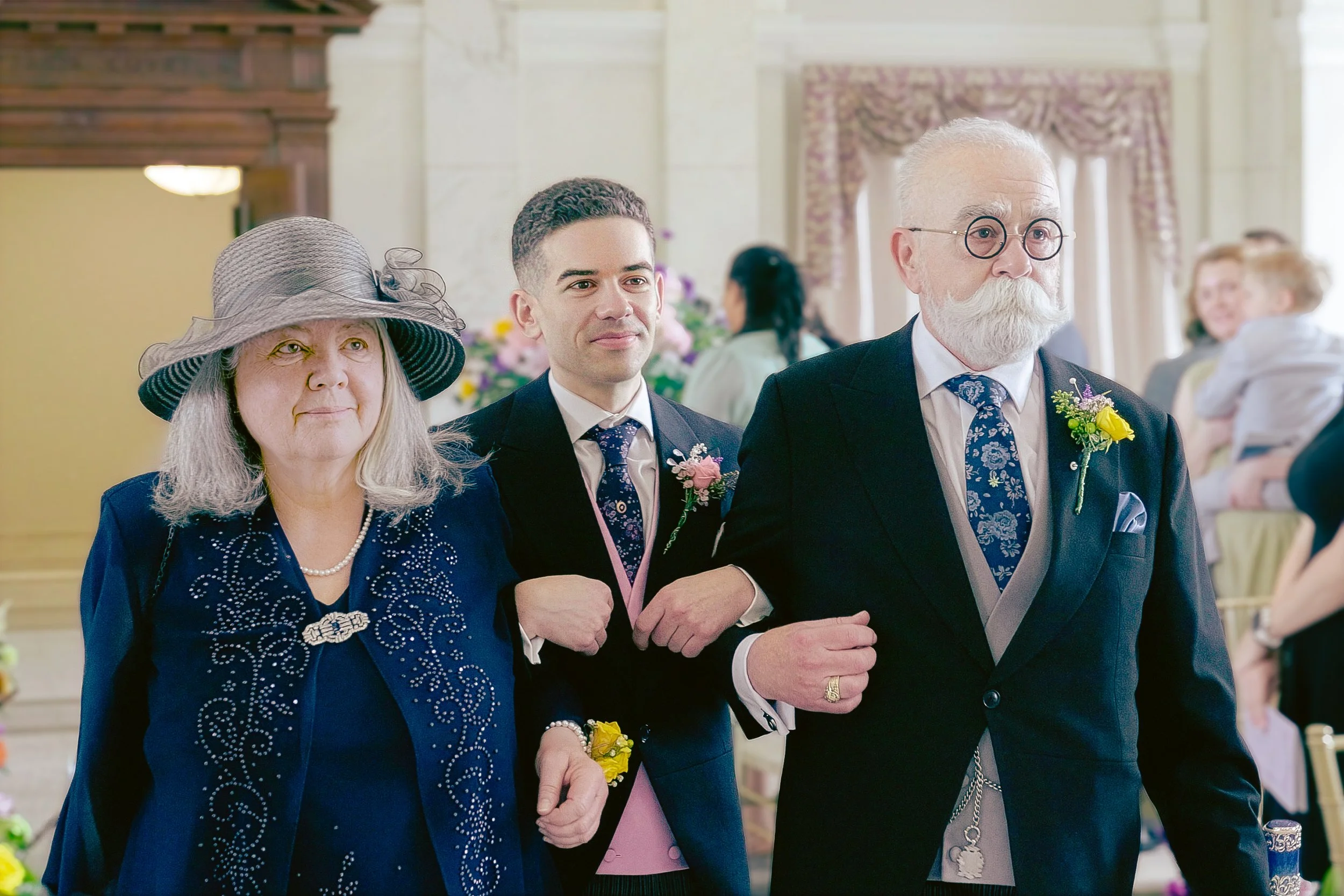 Three people, a woman with a hat, a young man, and an older man with glasses, are dressed in formal attire and holding hands at a wedding ceremony. They are standing in a decorated indoor venue with floral arrangements.