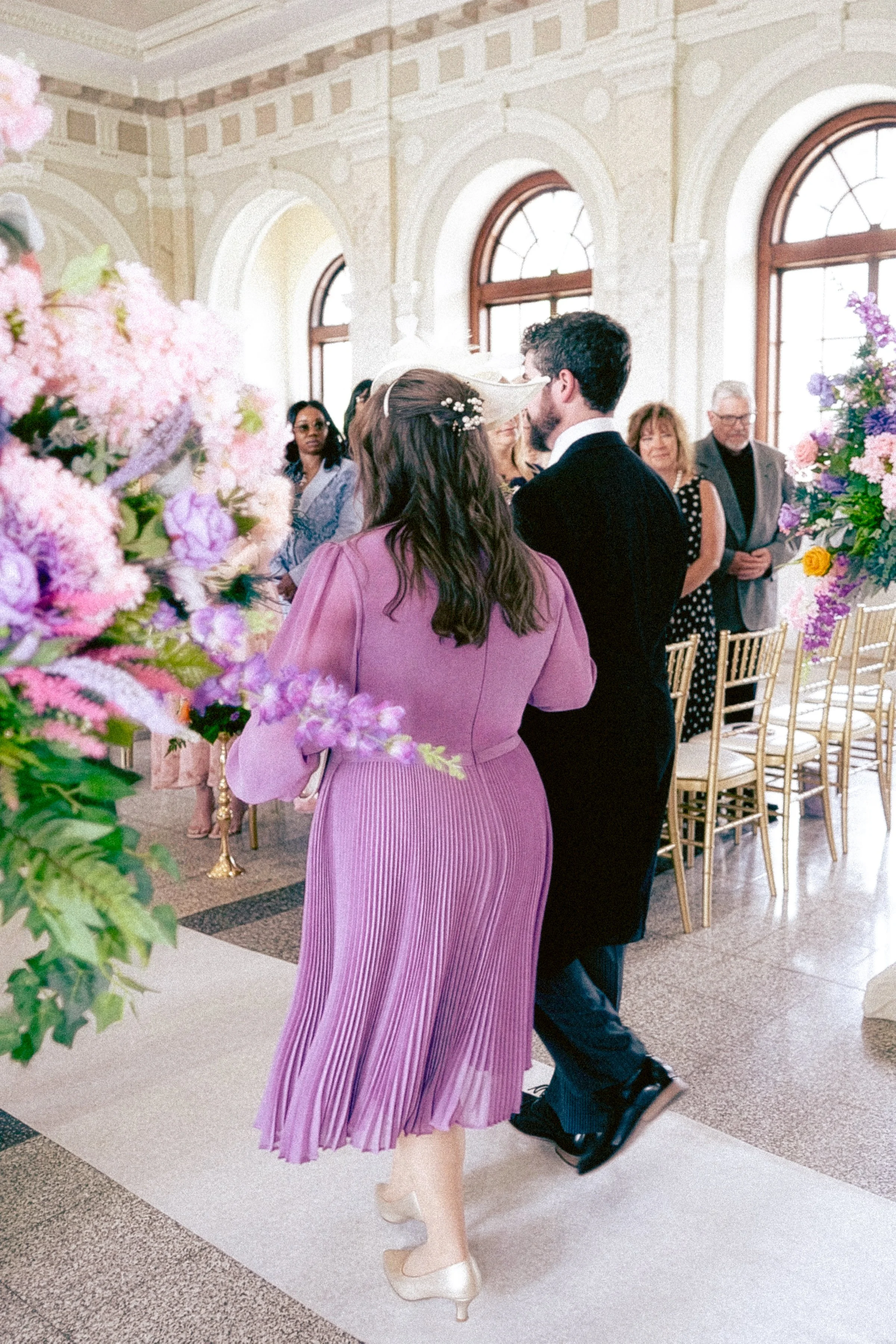 Couple dancing at a wedding reception, surrounded by floral arrangements and seated guests in a bright, elegant venue with large arched windows.