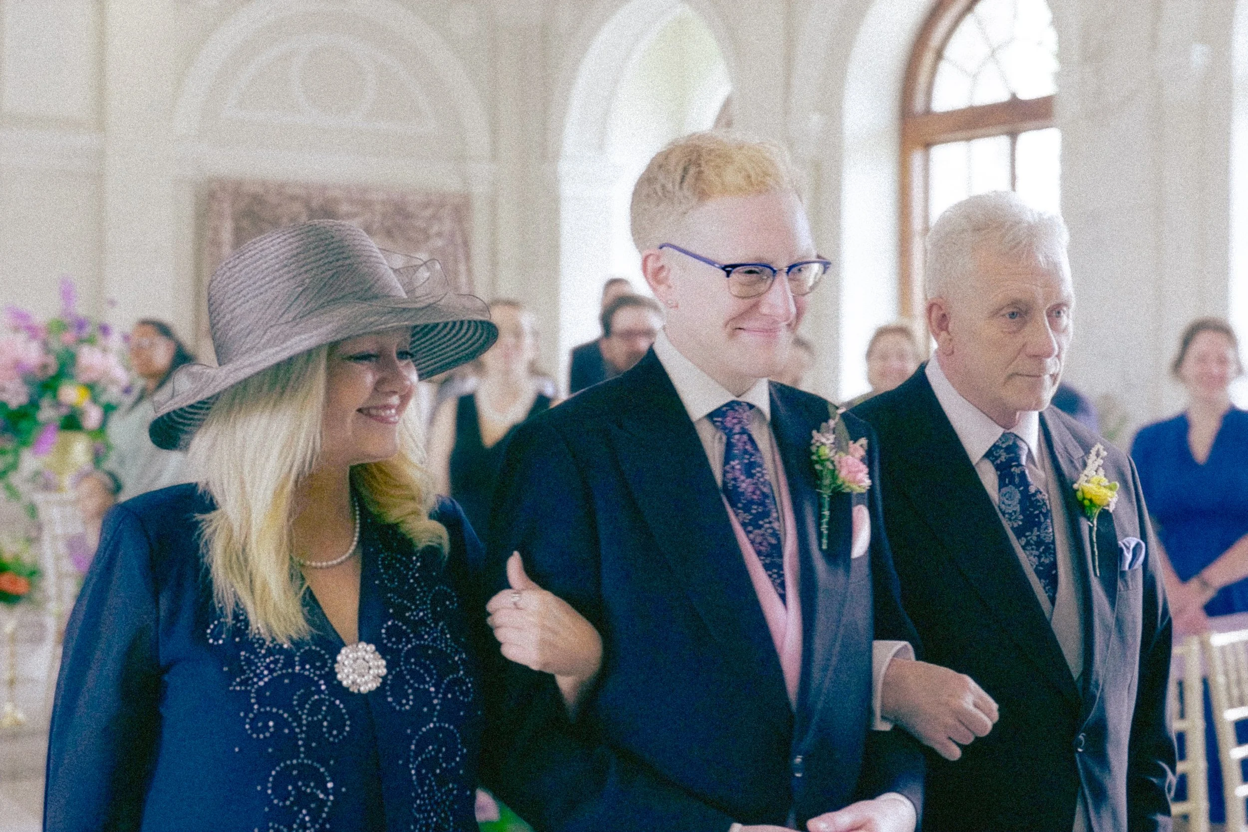 Group of formally dressed people attending a wedding ceremony in a decorated hall, with some holding hands or embracing.