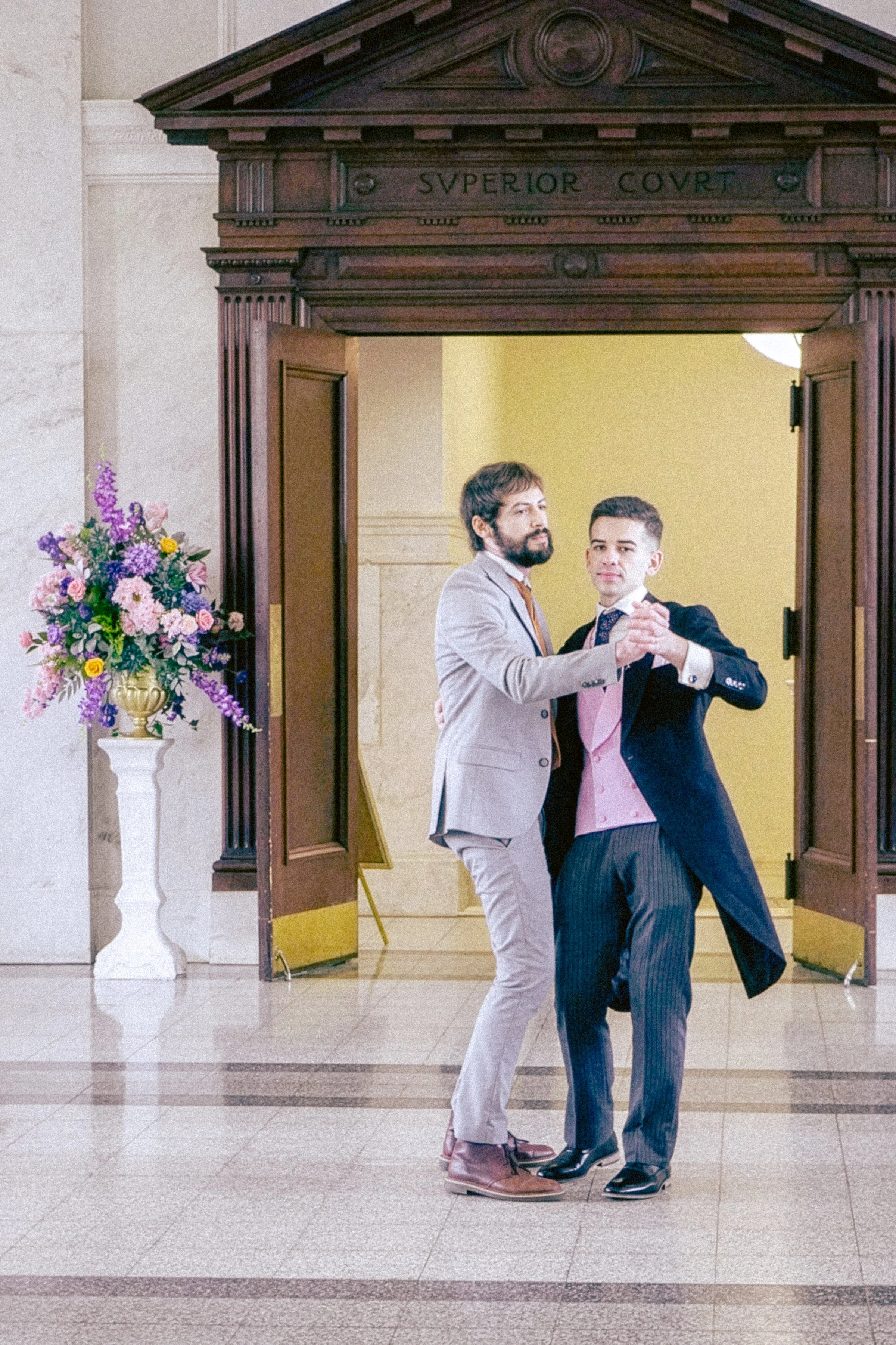 Two men dancing the tango inside a building with a wooden entrance labeled 'Superior Court.' One man wears a beige suit with brown shoes, and the other wears a dark suit with black shoes. They hold each other as they dance.