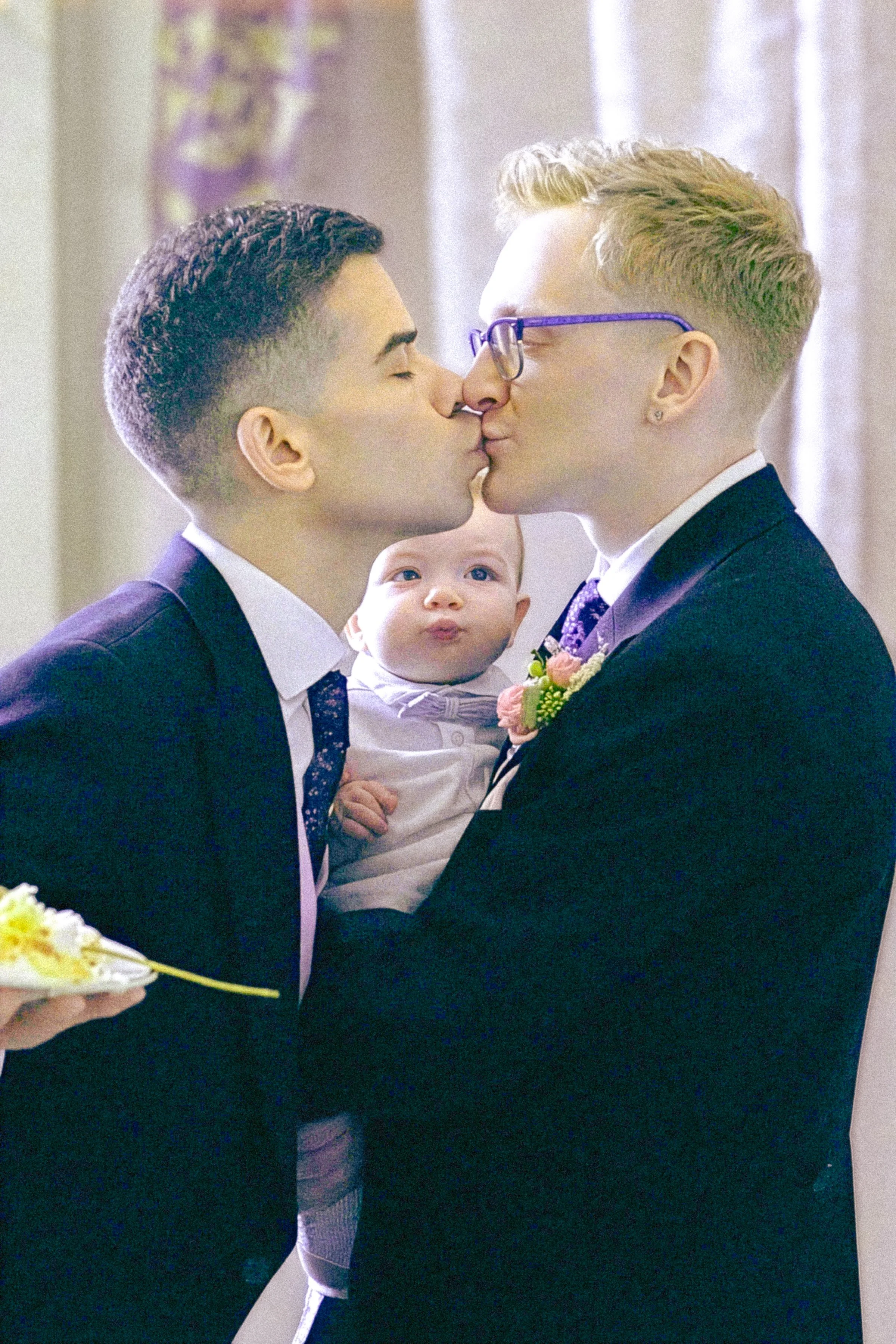 Two men in suits sharing a kiss on the lips, with a small child between them observing the moment. The man on the right wears glasses and has blonde hair, while the man on the left has dark hair.