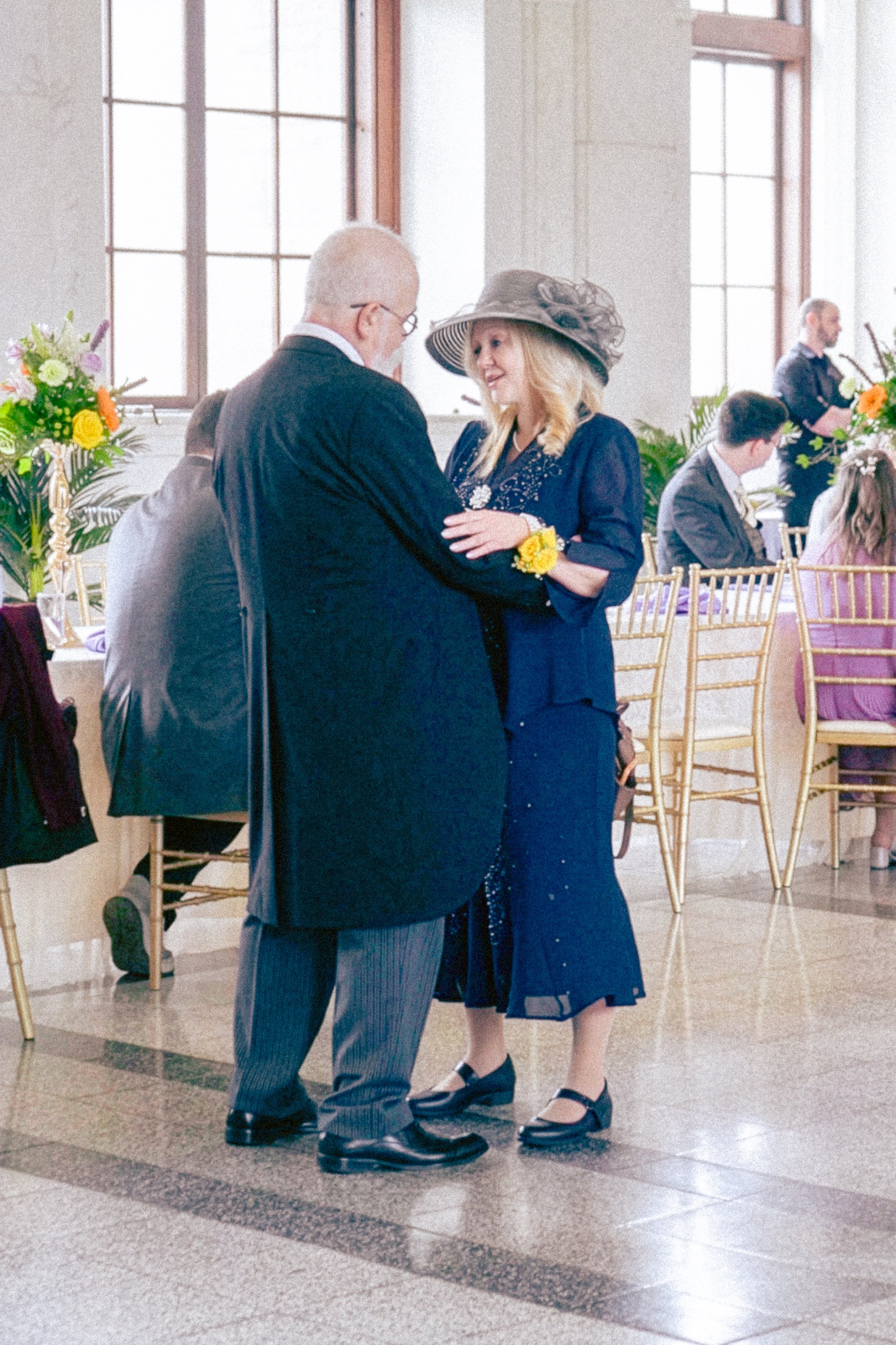 A man and a woman dancing together at a formal event, with the woman wearing a large hat and the man wearing a suit, inside a decorated room with tables and chairs, and other guests in the background.