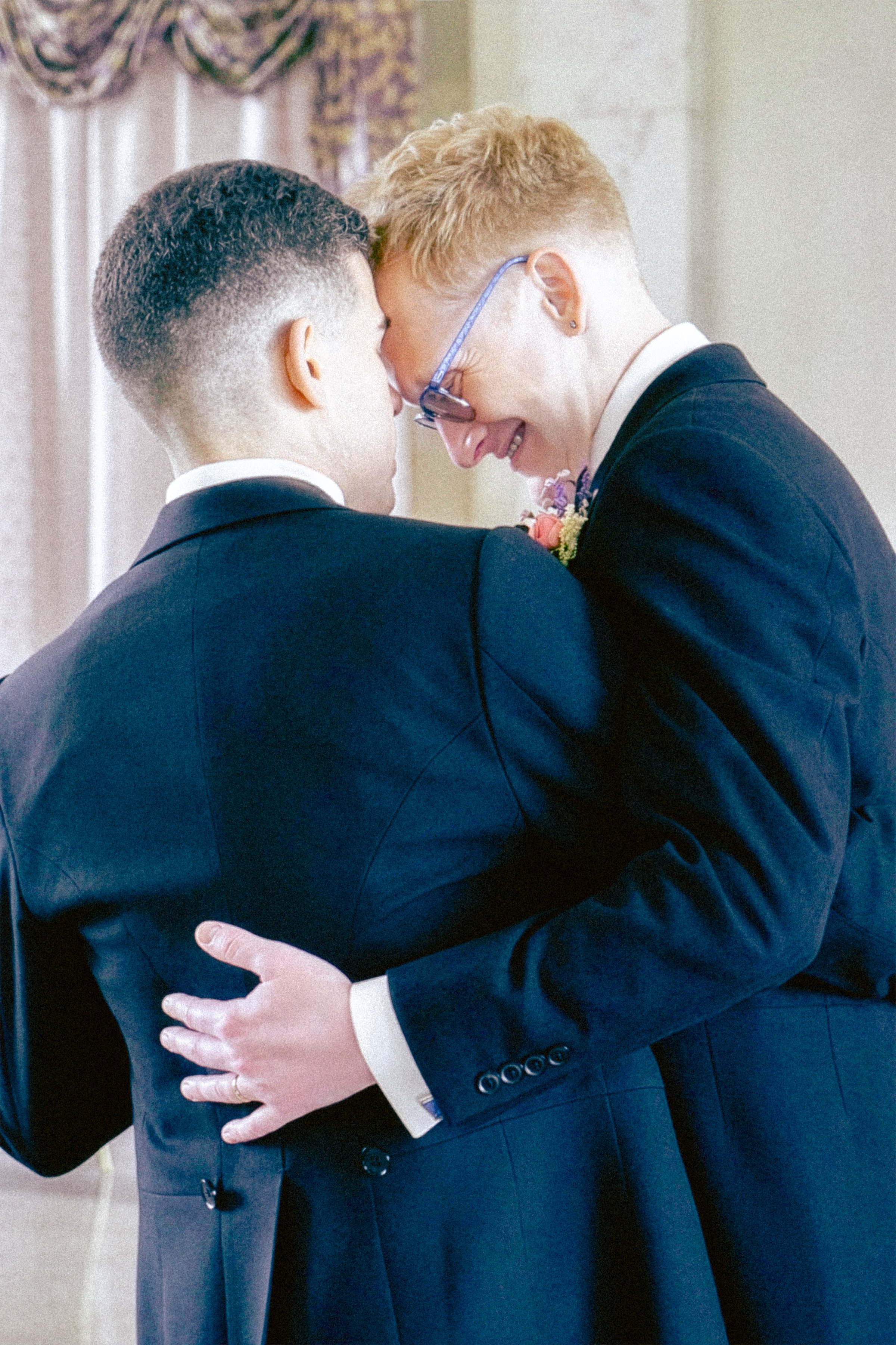 Two people, dressed in formal suits, are embracing in a close hug, with their foreheads touching and eyes closed.