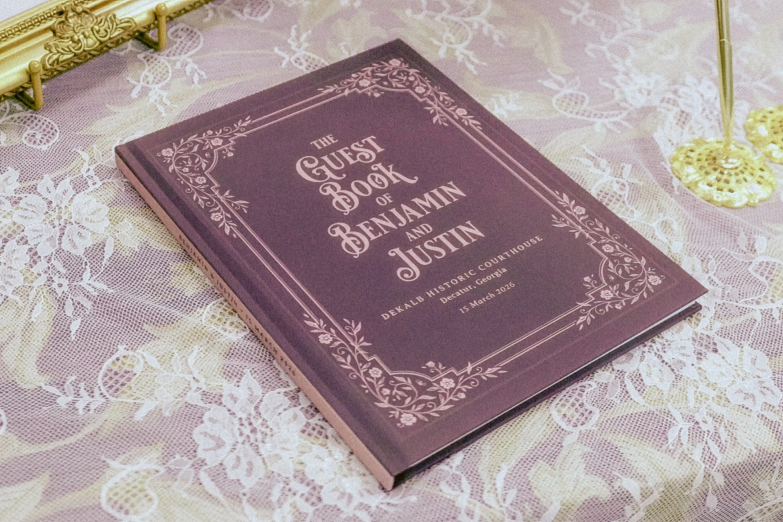 A purple book titled 'The Guest Book of Benjamin and Austin' lies on a lace tablecloth with a gold-colored decorative table beside it. A small yellow decorative item is on the right side of the image.