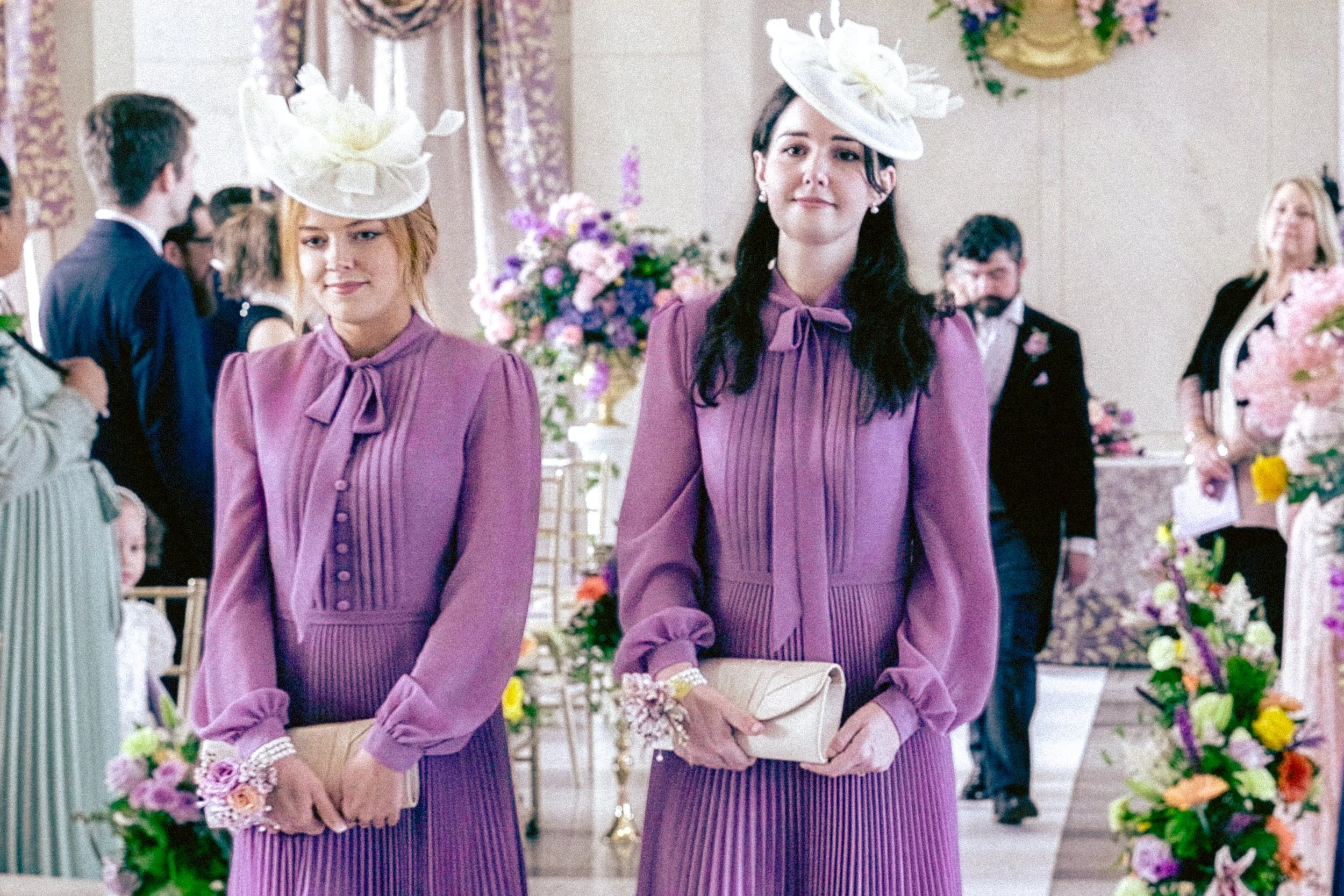 Two women wearing matching purple dresses and large white floral hats stand in a decorated indoor setting, likely a wedding or special event, with other guests and flower arrangements in the background.