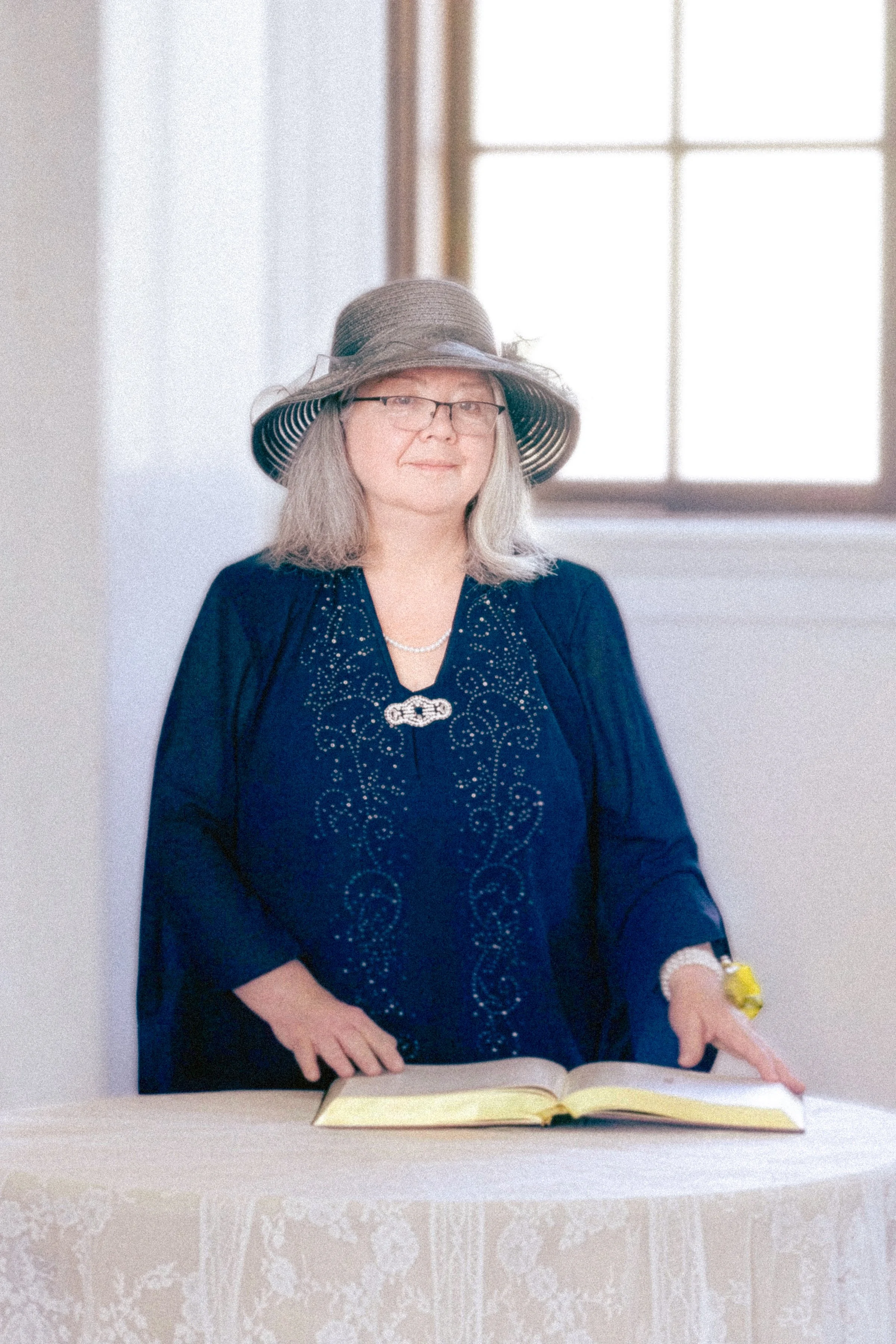 A woman with gray hair, glasses, and a large sun hat stands behind a table with a book open in front of her, in front of a large window with multiple panes, in a well-lit room.