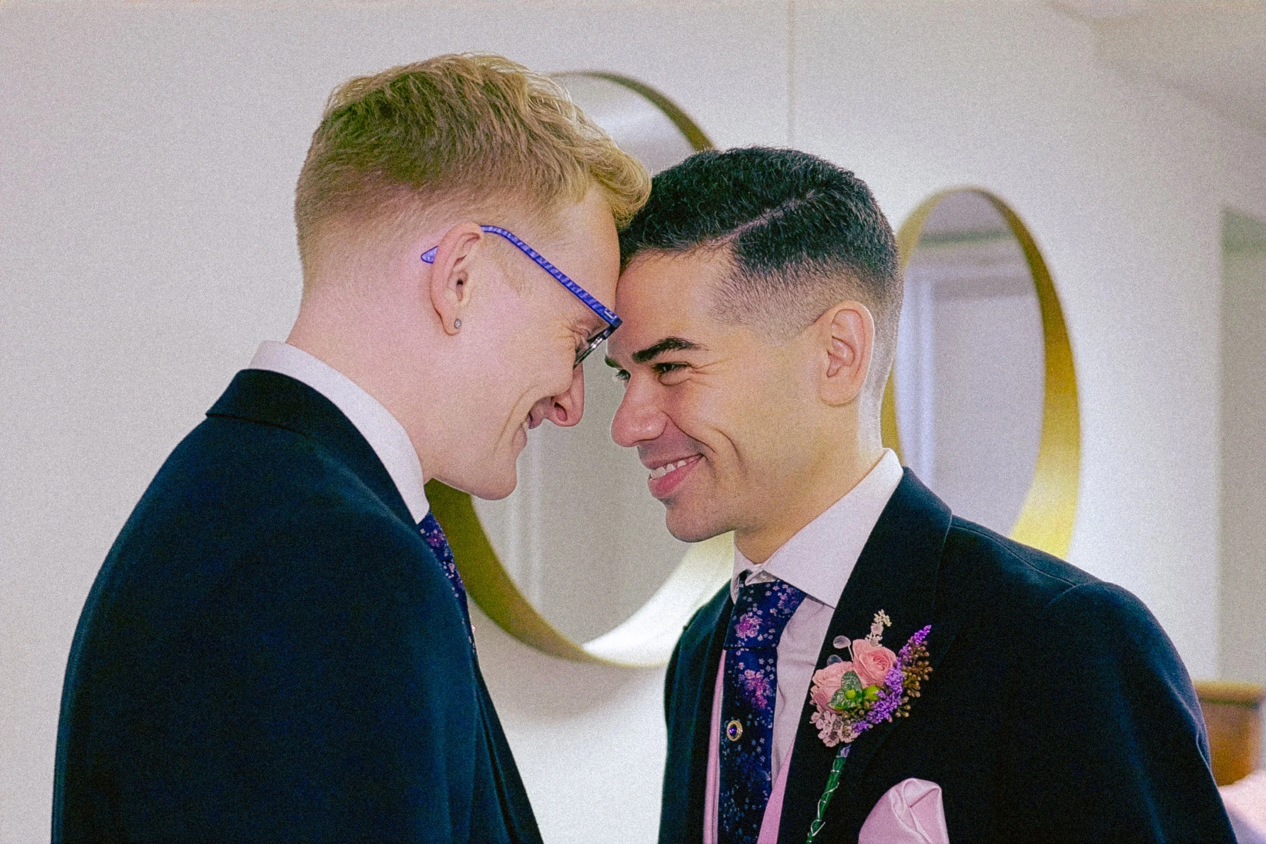 Two men dressed in tuxedos face each other closely, smiling, with their foreheads touching. One has blond hair and glasses, the other has dark hair and a boutonniere with pink and purple flowers. In the background, there are round mirrors on the wall.