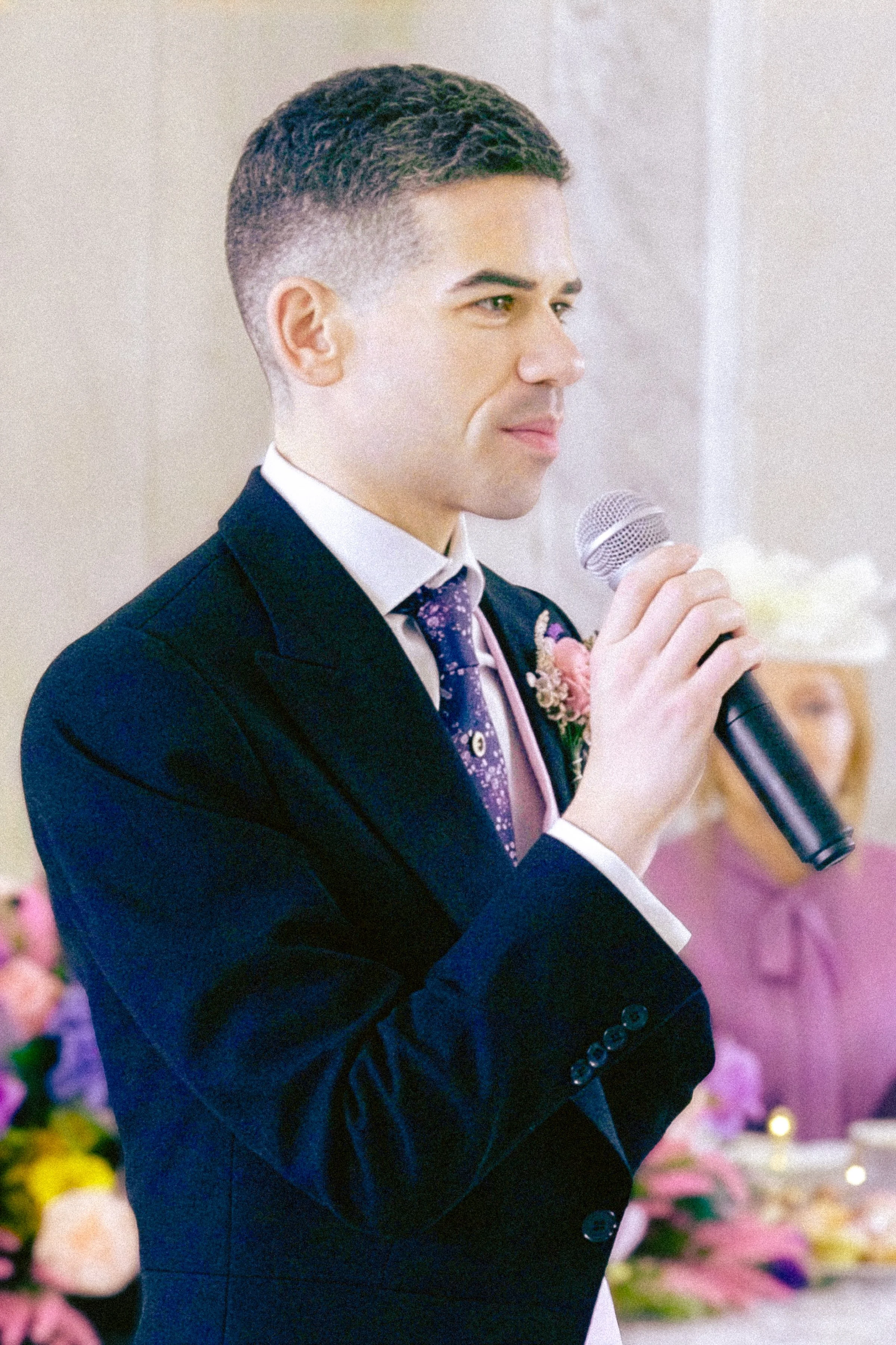 A young man in a black suit holding a microphone at a celebration or formal event. The background includes colorful flowers, indicating a festive atmosphere.