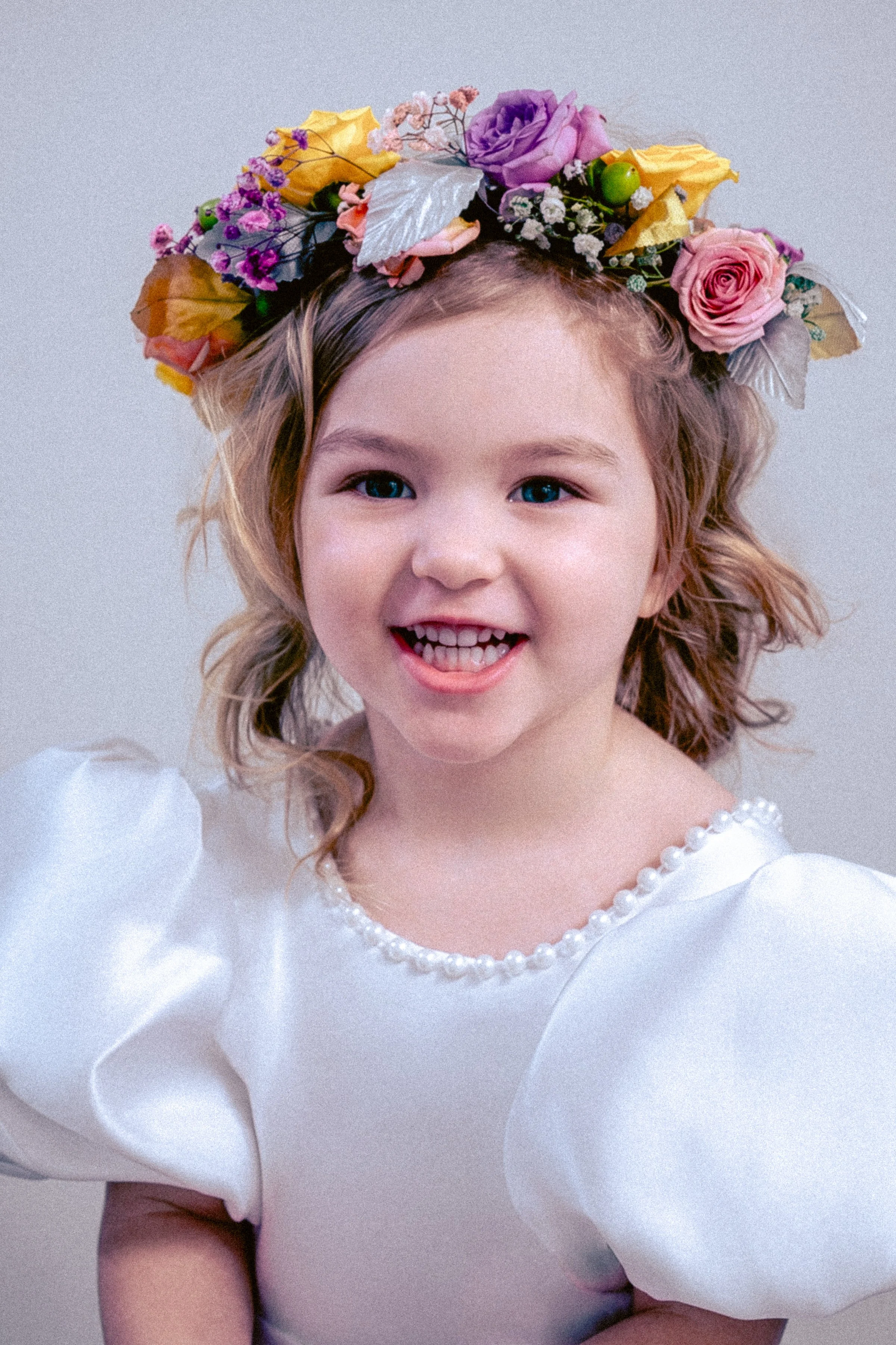 A young girl with blonde hair and blue eyes wearing a white dress with puffy sleeves, decorated with pearls around the neckline, and a colorful flower crown on her head.