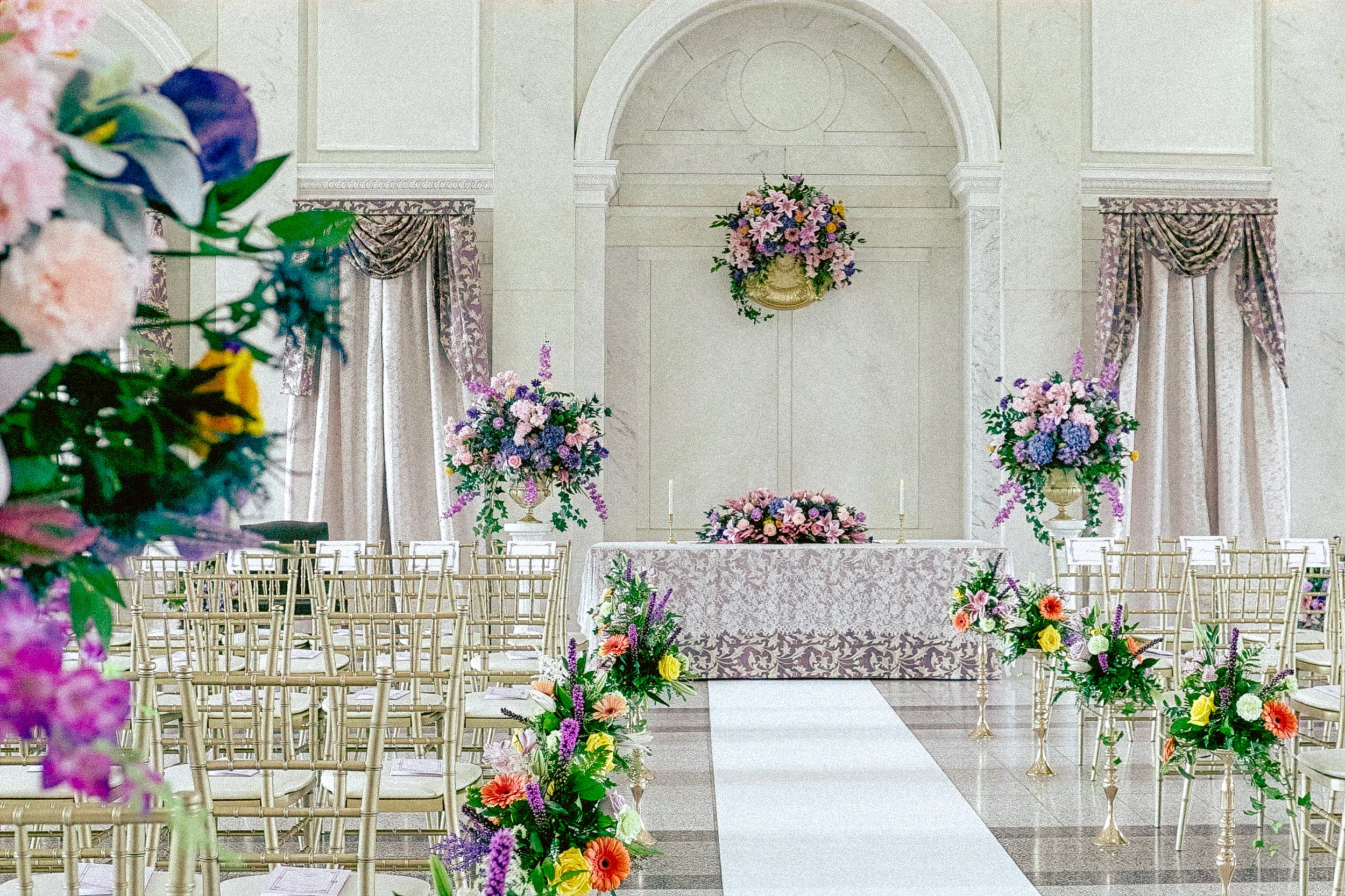 Elegant wedding ceremony setup with flower arrangements, white chairs, and a decorated altar in a bright, spacious hall.