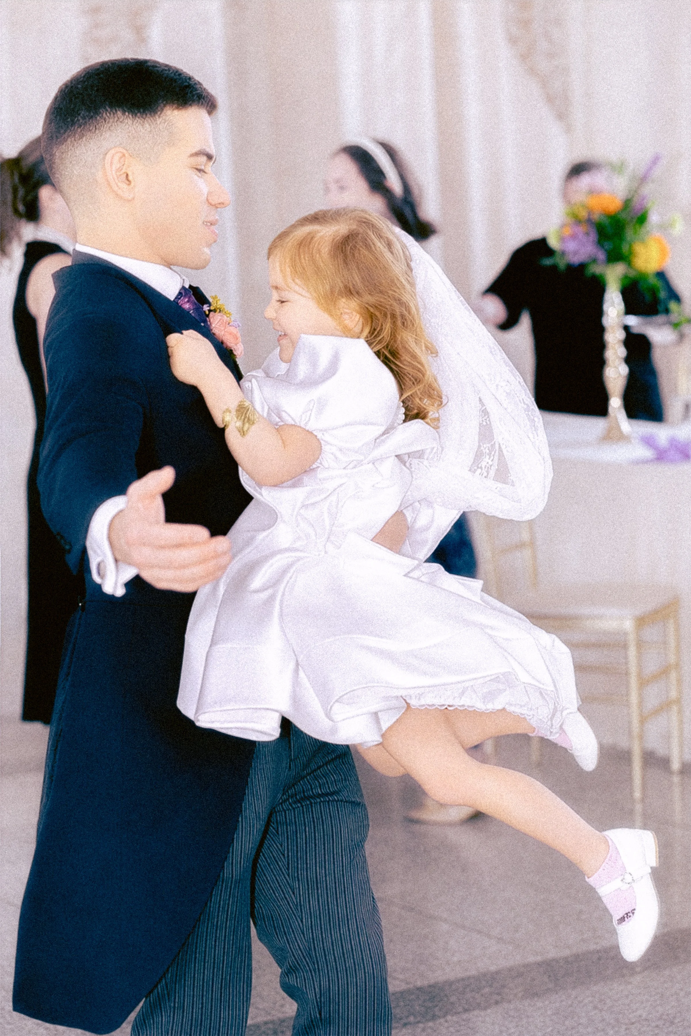 A man in a suit dancing with a young girl in a white dress at a formal event, possibly a wedding reception, with other guests and floral arrangements visible in the background.