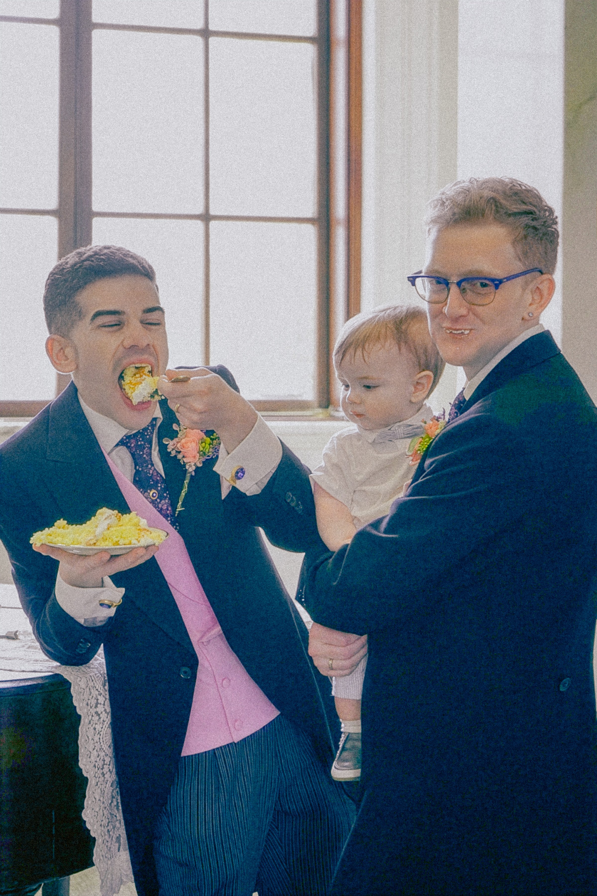 Two men dressed in formal suits, one feeding cake to the other, with a child standing with them. The scene appears to be a celebration, possibly a wedding.