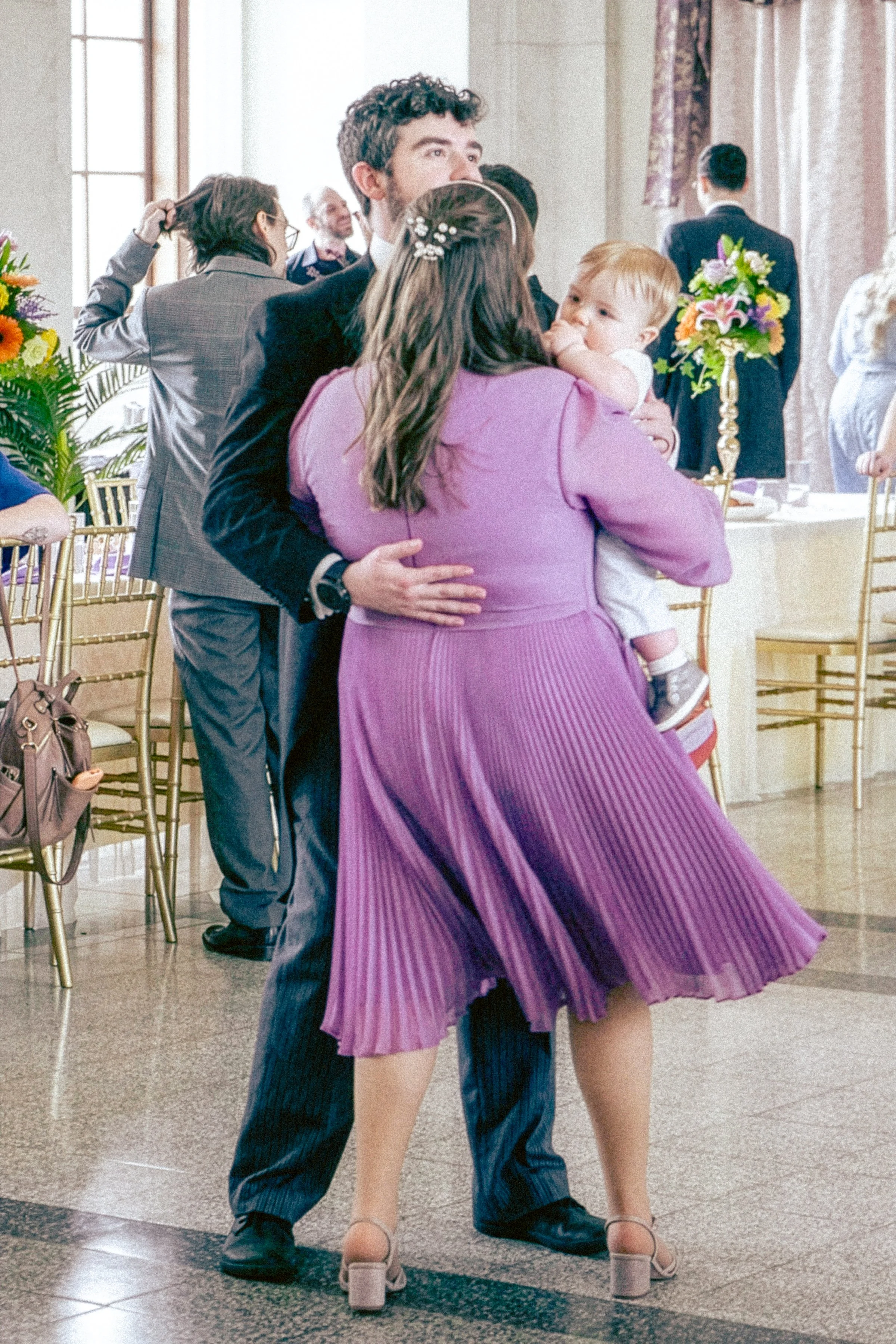 A man, woman, and child dancing closely at a formal indoor event with floral centerpieces and gold chairs.