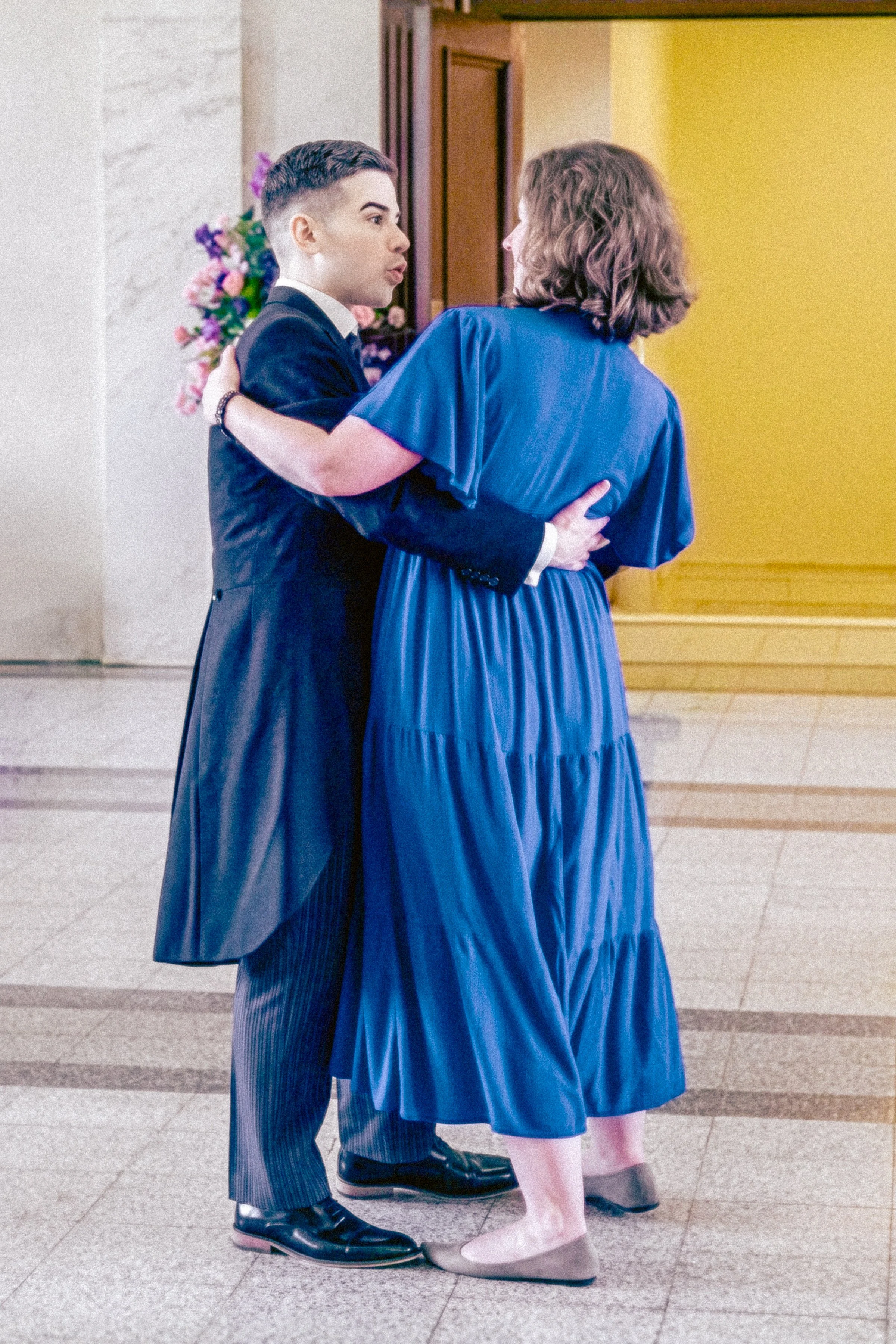 A young man and woman are dancing closely together indoors. The young man, dressed in a formal dark suit and striped pants, is holding the woman by her waist. The woman is wearing a long blue dress and beige shoes, with her arm around the man's shoul