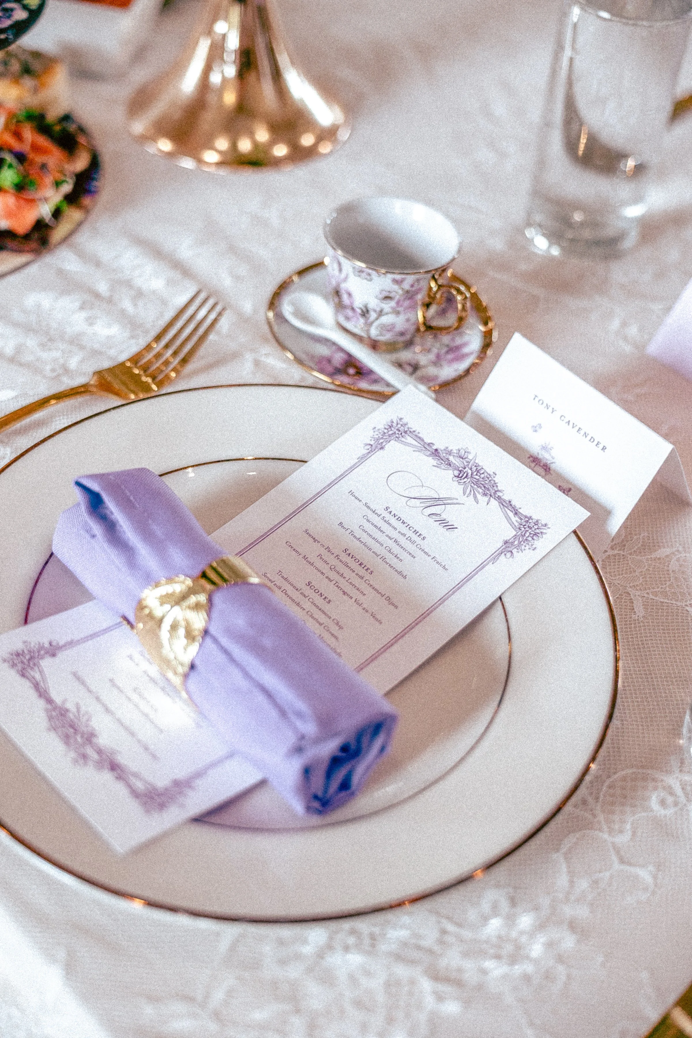 Close-up of a table setting featuring a white and gold-rimmed plate, a lavender napkin, a purple box with a gold seal, a decorative teacup with floral pattern on saucer, a menu card, and a gold-colored fork with a glass of water in the background.