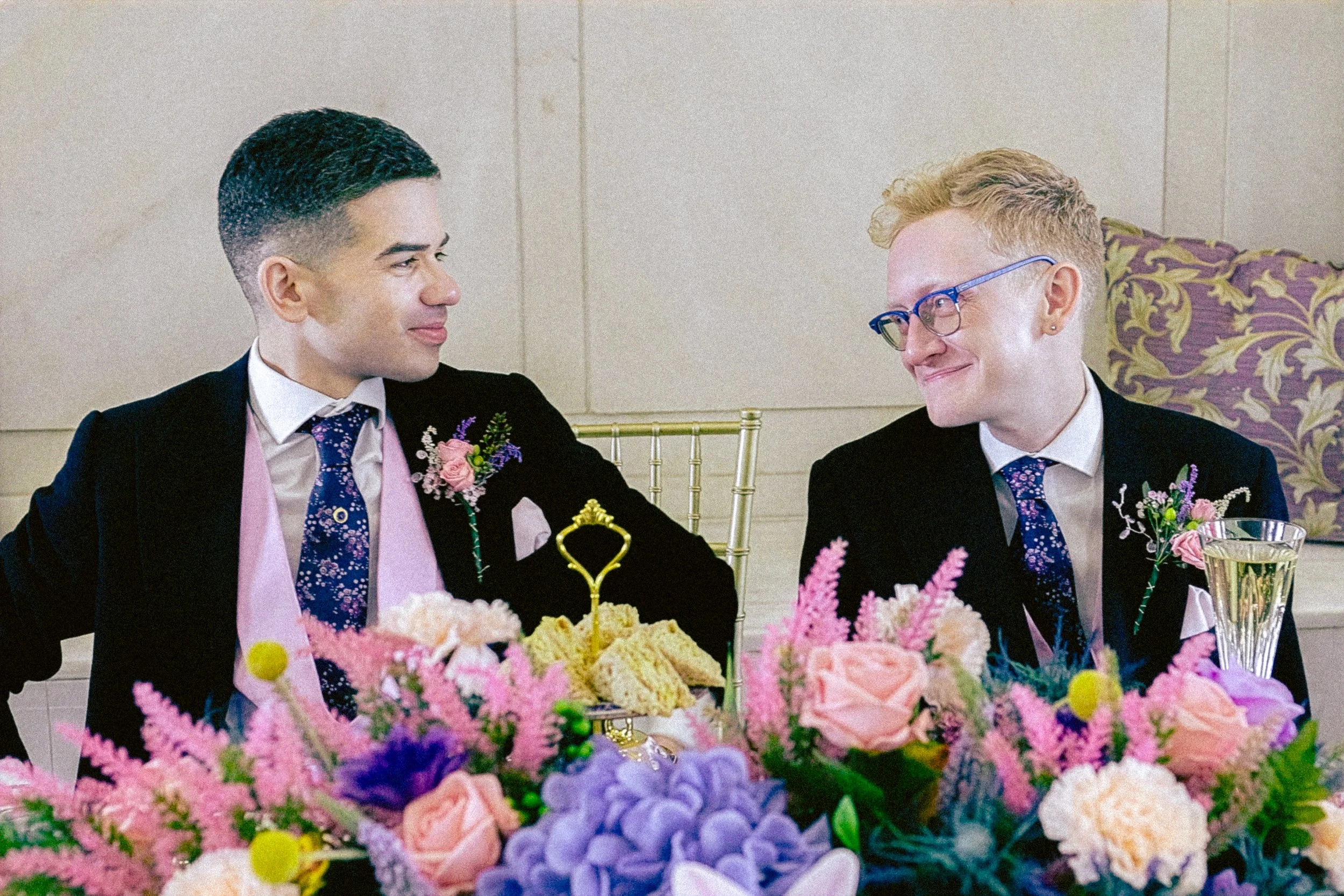 Two individuals dressed in formal attire sitting at a table with a floral centerpiece, smiling at each other during a celebration or event.