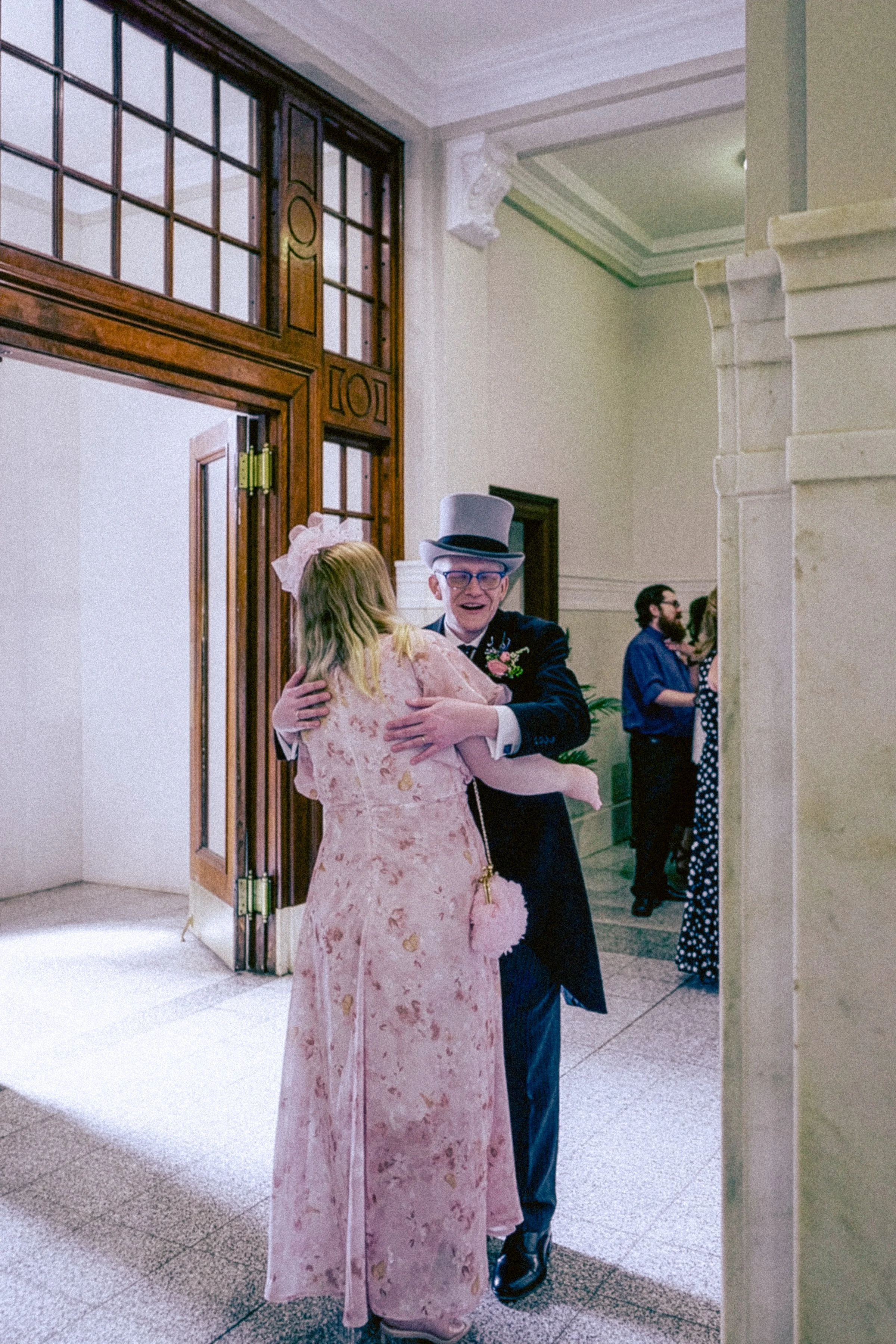 A man in a suit and a woman in a pink dress hugging each other at a formal event, with other people in the background inside a building with wood and stone architectural details.