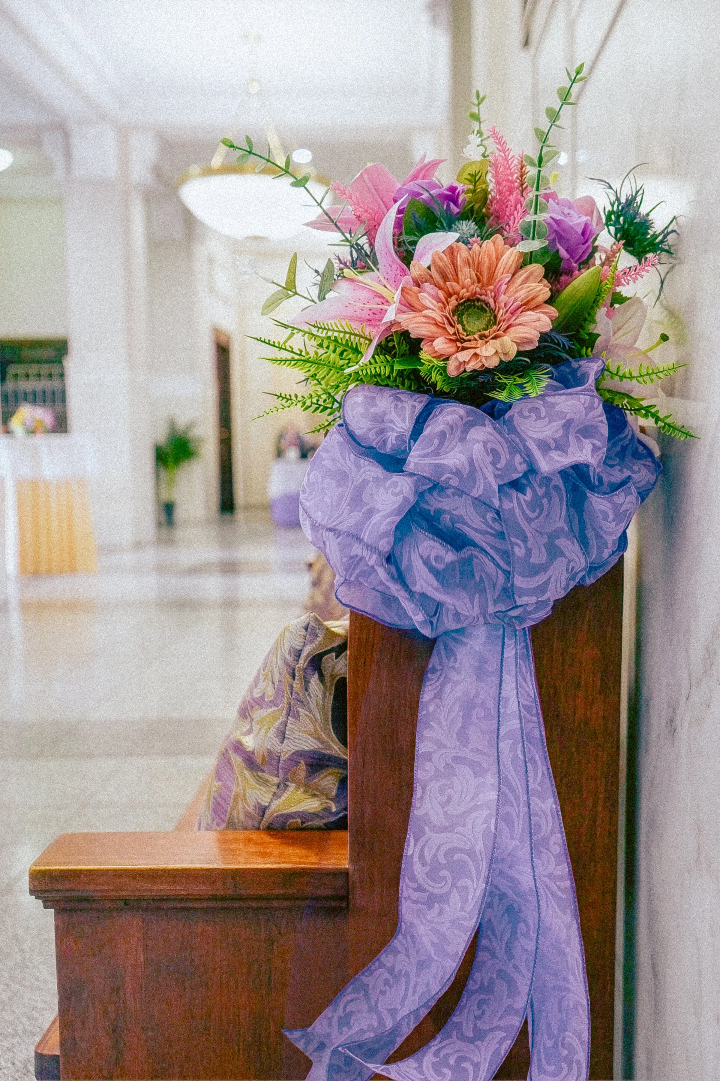 A bouquet of mixed flowers, including pink lilies, orange gerbera daisies, purple roses, and greenery, wrapped in purple patterned paper, on a wooden table in an indoor setting.