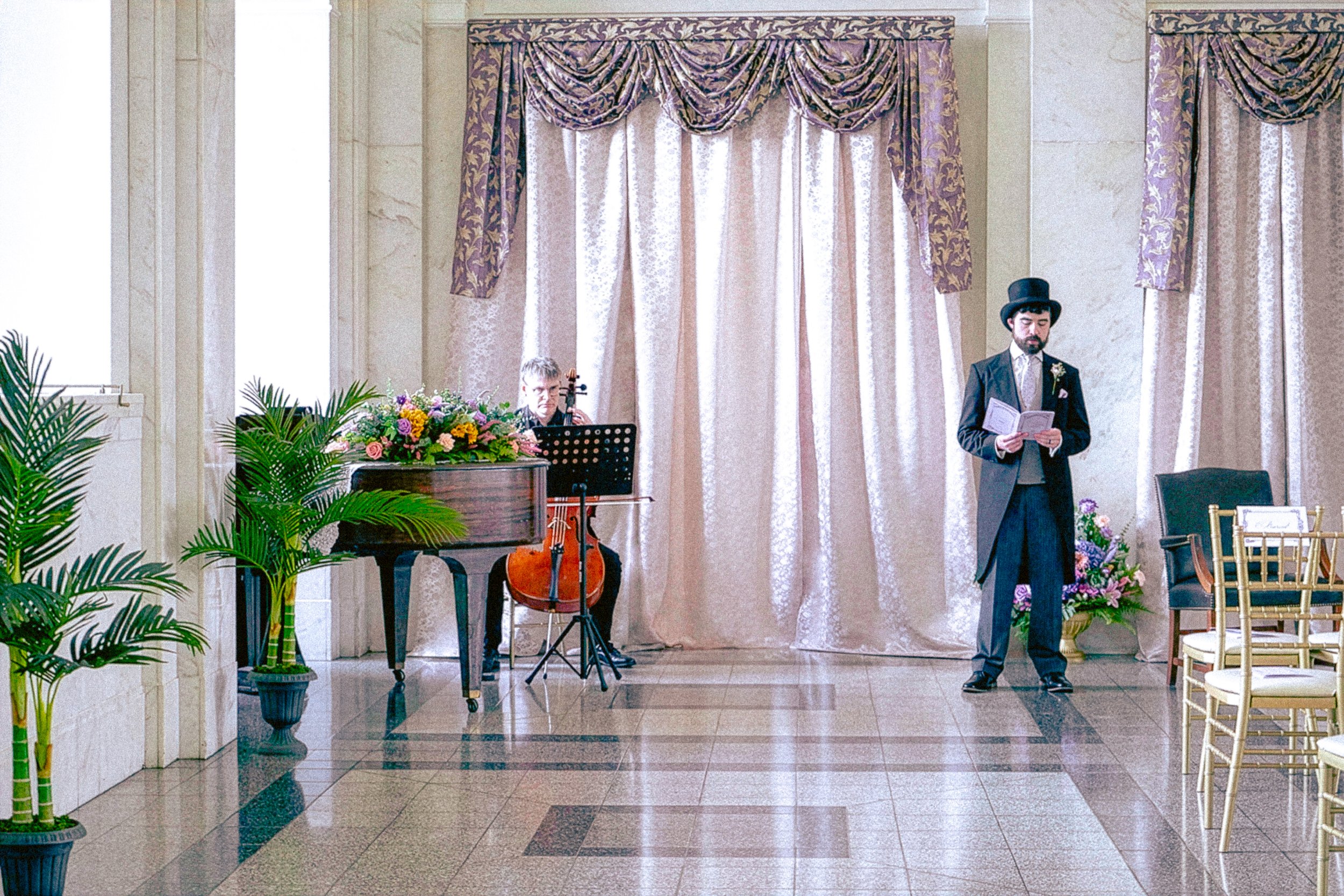 Man in Victorian attire reading at a wedding ceremony, with musician playing the cello and floral arrangements in a decorated indoor venue.