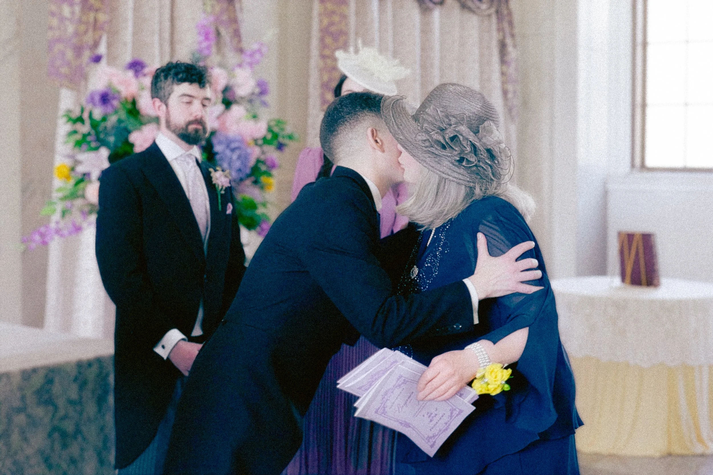A woman wearing a large gray hat and blue dress embraces a man in a dark suit at a formal event. The woman is holding pink papers and yellow flowers, and a floral arrangement is visible in the background.