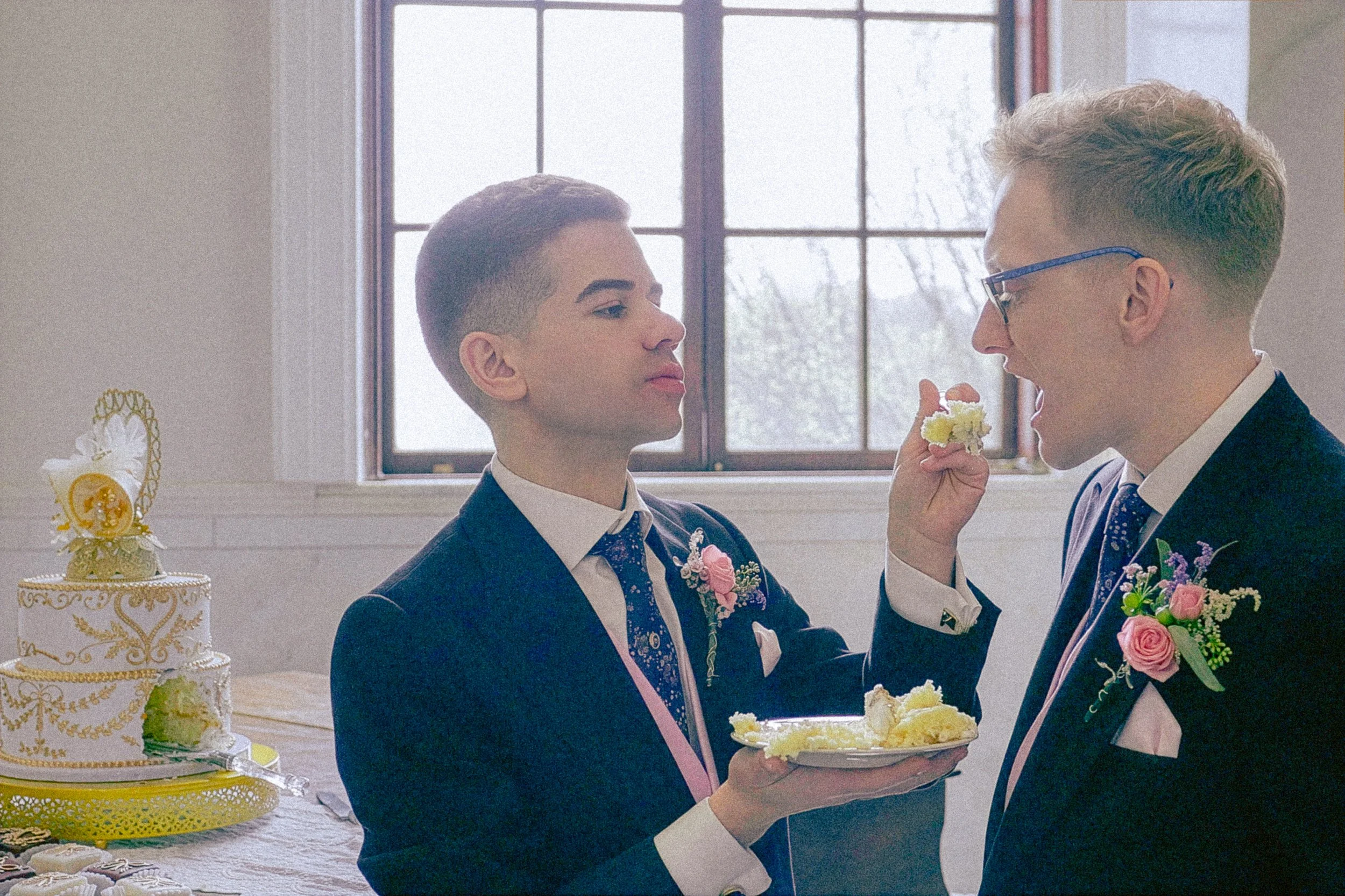 Two men in formal suits sharing a piece of wedding cake at a celebration, with a decorated wedding cake on the table beside them and a large window in the background.