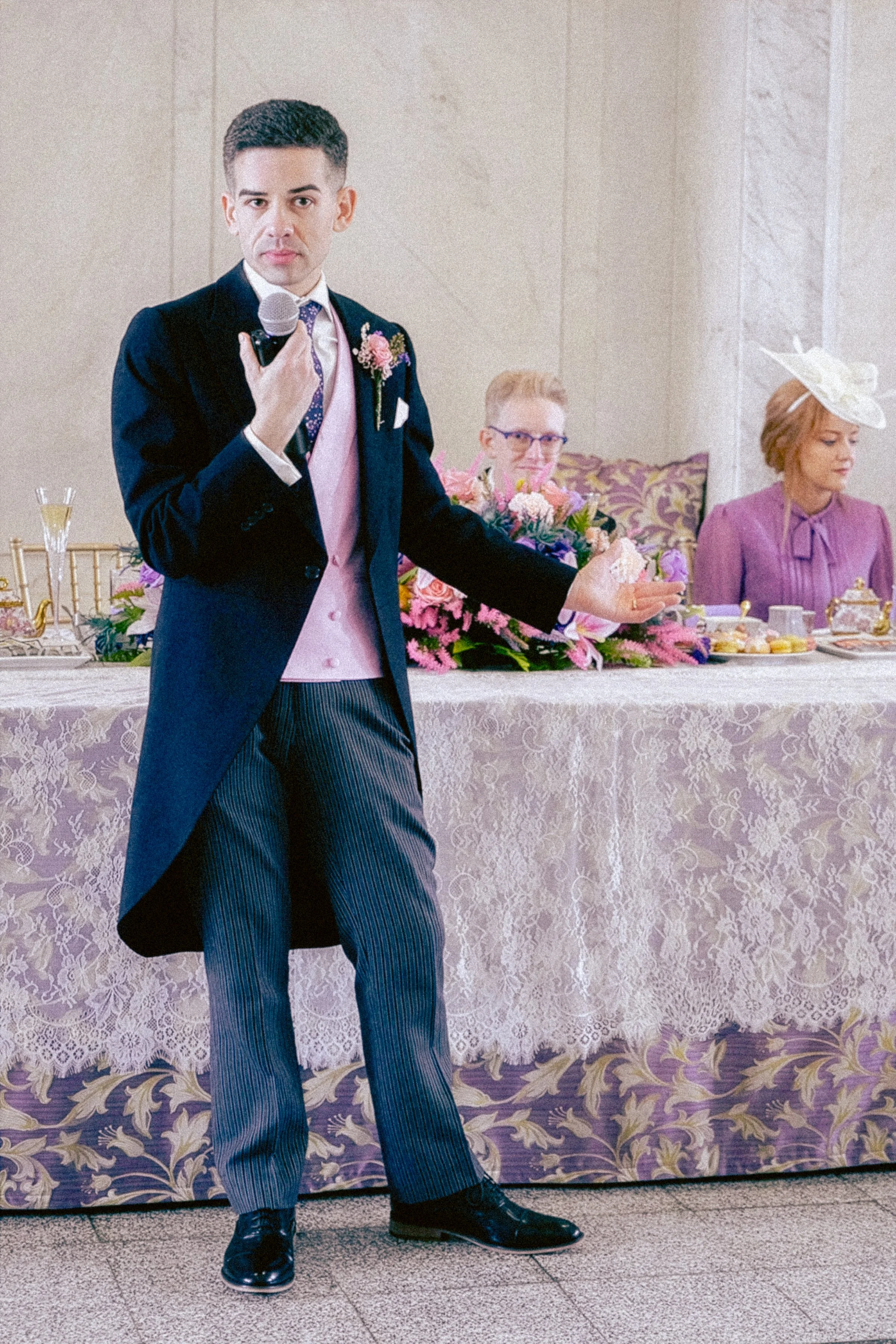 Man in black tuxedo with purple vest and tie holding a microphone at a wedding reception. Two women are seated behind him, one with floral bouquet and the other wearing a purple dress and a large white hat.