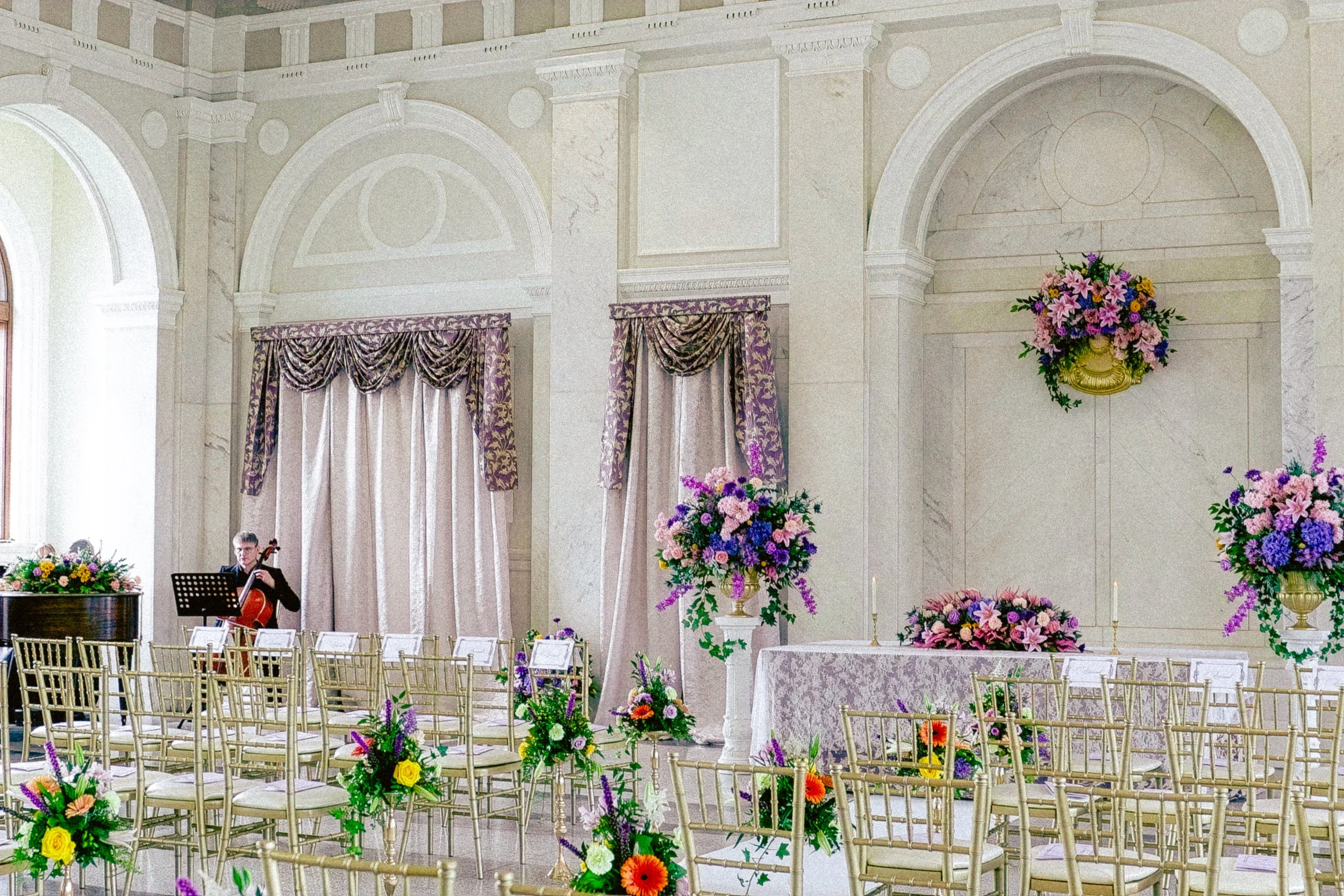 Elegant wedding ceremony setup in a large hall with floral arrangements, gold chairs, and a musician playing a cello.