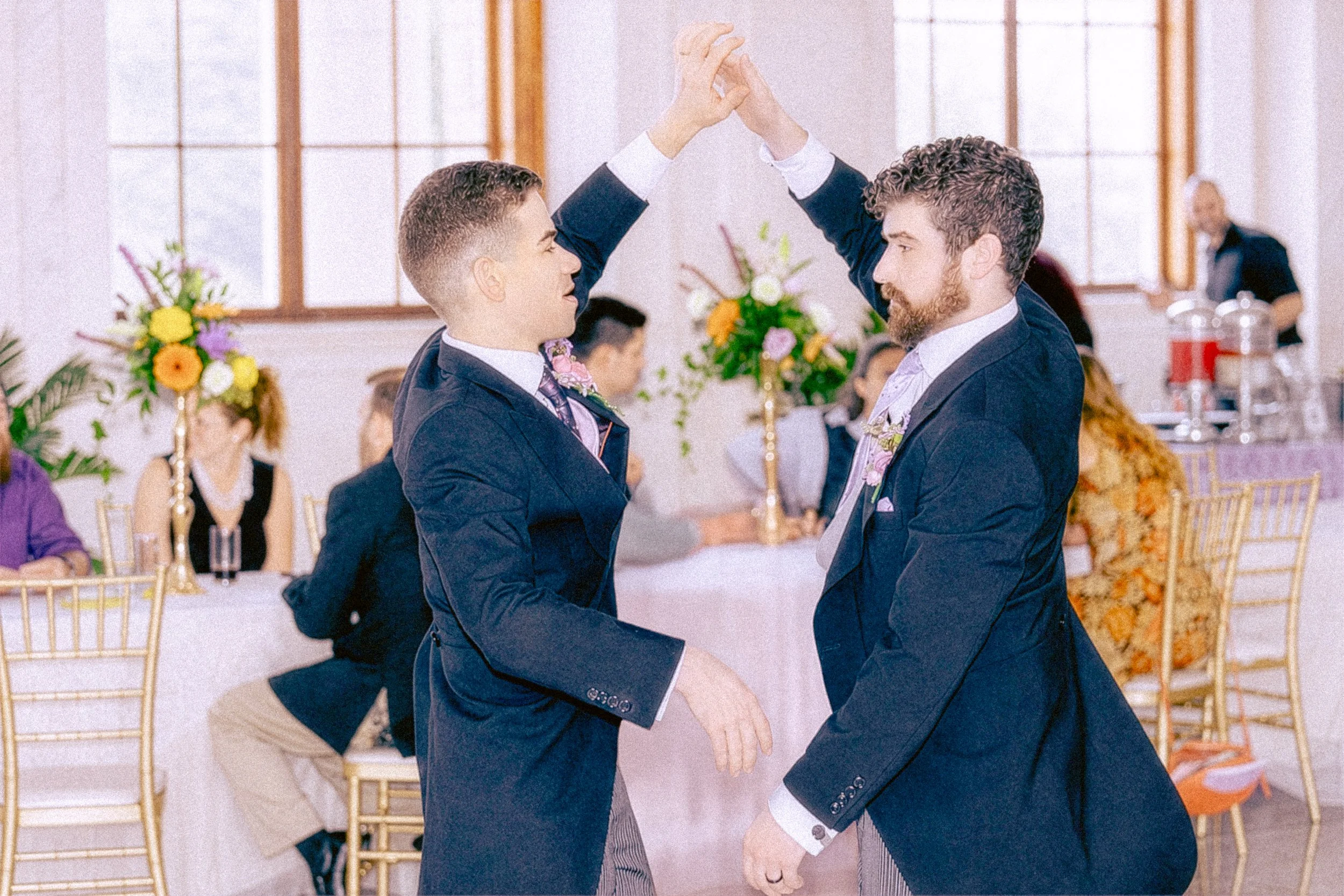 Two men in formal attire dancing at a wedding reception, holding hands and smiling at each other, with guests and floral arrangements in the background.