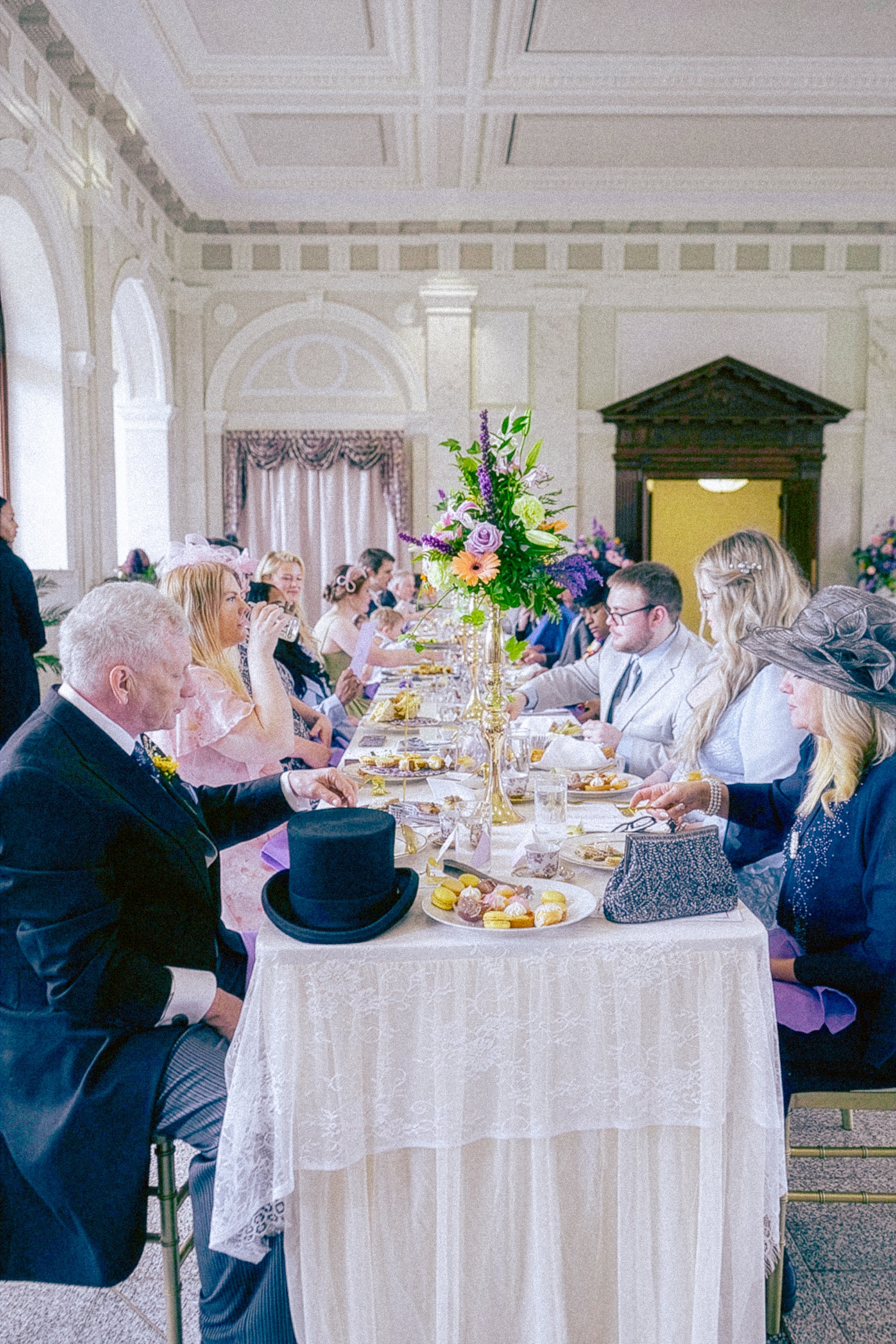 Guests seated at a long table during a formal event, with floral centerpieces, dressed in elegant attire, including a man wearing a tuxedo with a top hat, and women wearing dresses and hats, in a decorated banquet hall.