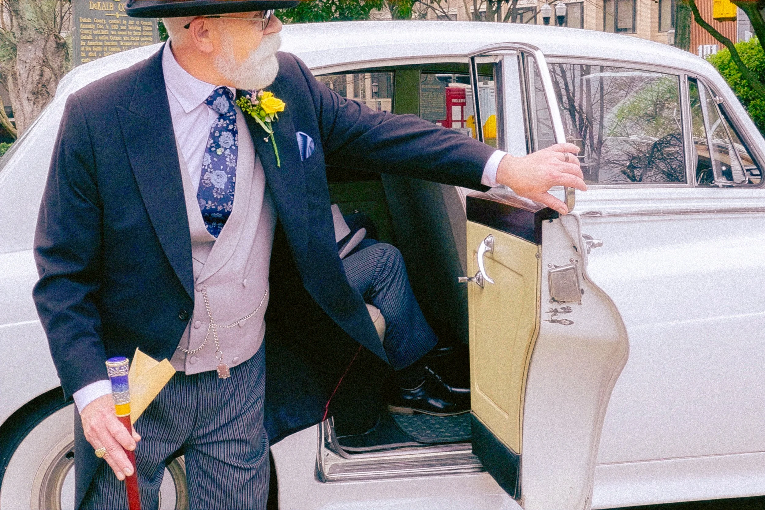 A man dressed in a formal suit with a boutonniere, wearing a hat and glasses, is stepping out of a vintage yellow and cream-colored car, holding a cane in his right hand, on a city street with trees and buildings in the background.