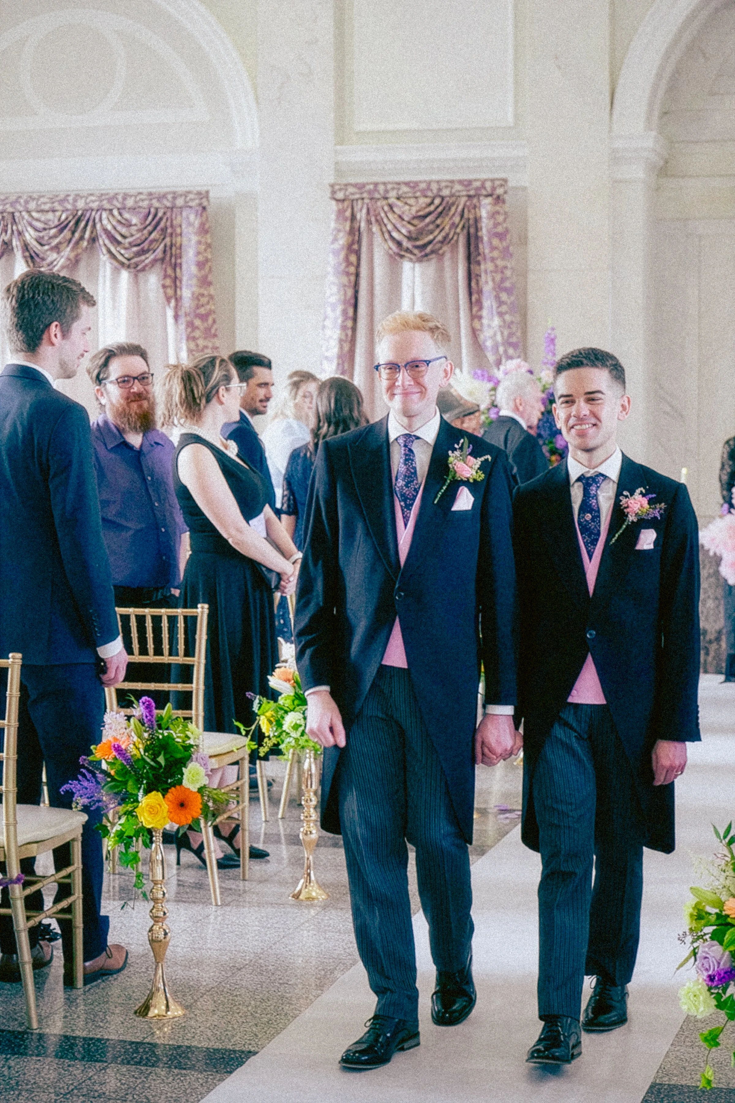 A group of people at a formal wedding ceremony. Two men in the foreground are holding hands and walking down the aisle, dressed in suits with boutonnieres. Guests are standing and watching, with some smiling. The room has elegant curtains, flowers, a