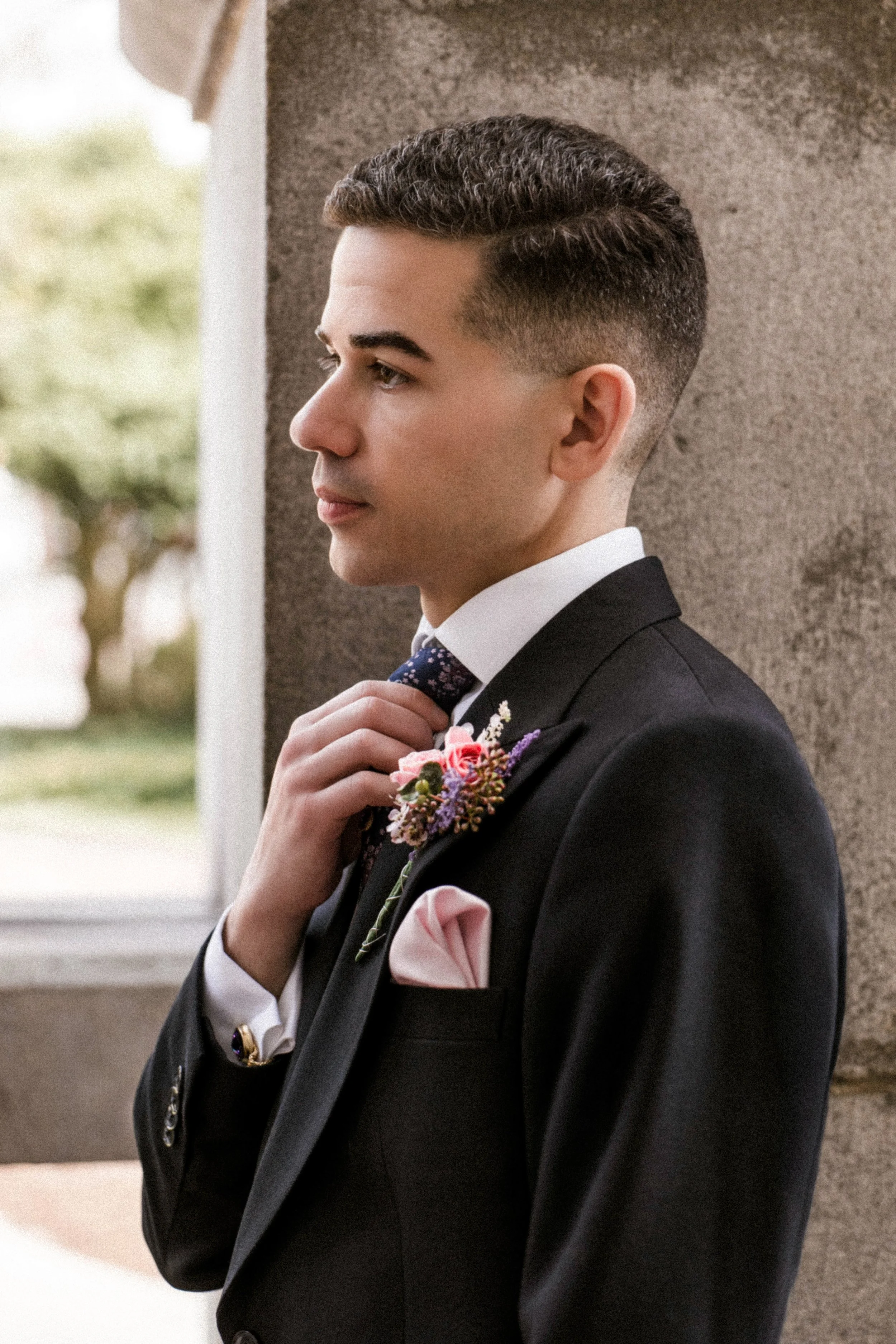 A young man in a tuxedo adjusting his bow tie, standing outdoors near a textured wall with trees in the background.