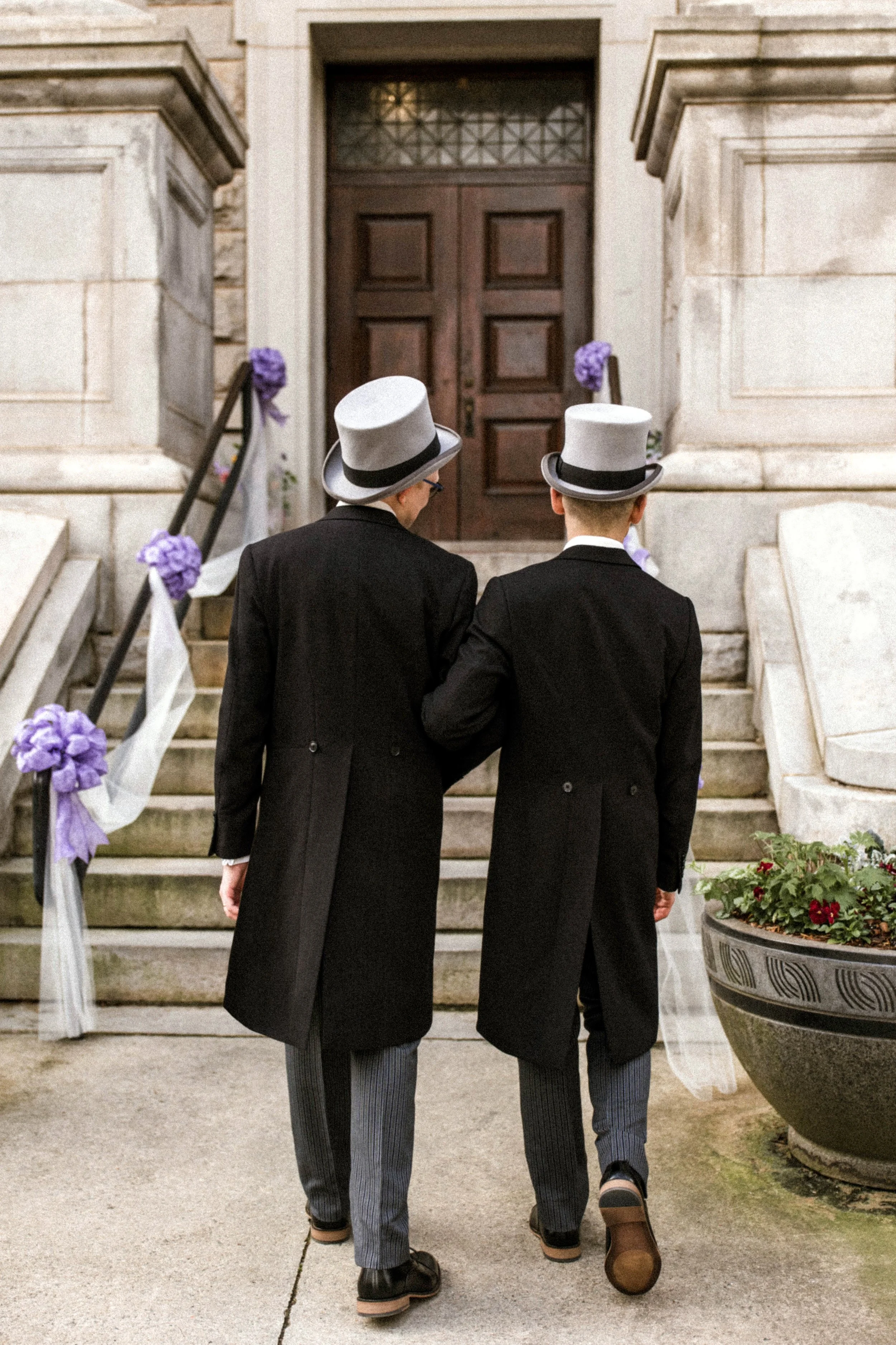 Two men in tuxedos with top hats walking up the steps of a building decorated with purple flowers for a wedding or special event.