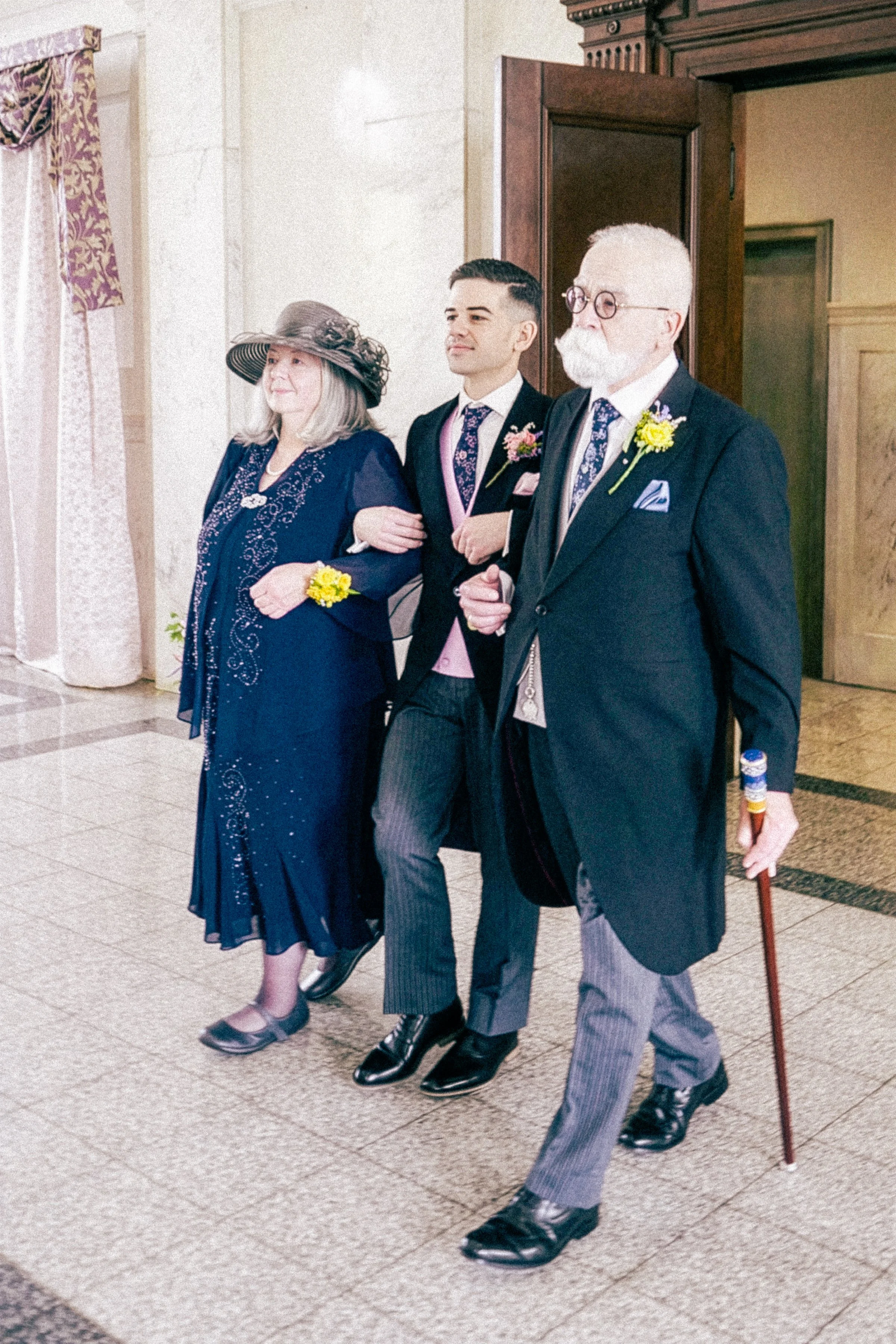 Three people dressed in formal attire are walking arm-in-arm in an indoor setting. The woman on the left is wearing a dark blue dress with embroidery, a large hat, and has a yellow boutonniere. The man in the middle is wearing a tuxedo with a pink wa