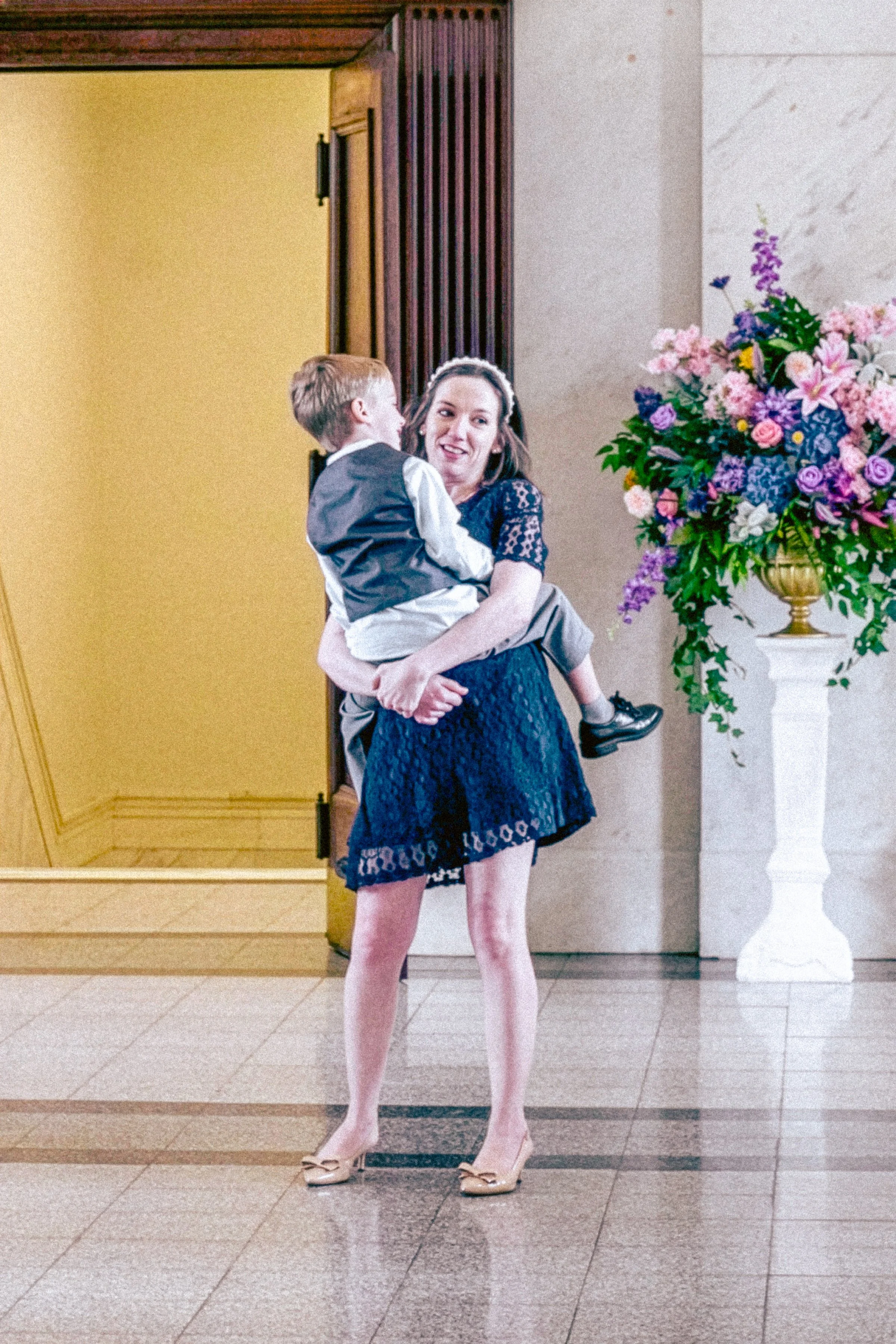 A woman holding a young boy inside a building with a large floral arrangement on a tall white pedestal nearby. The woman is smiling and wearing a navy blue dress, while the boy is dressed in a gray vest and white shirt. The background includes a yell