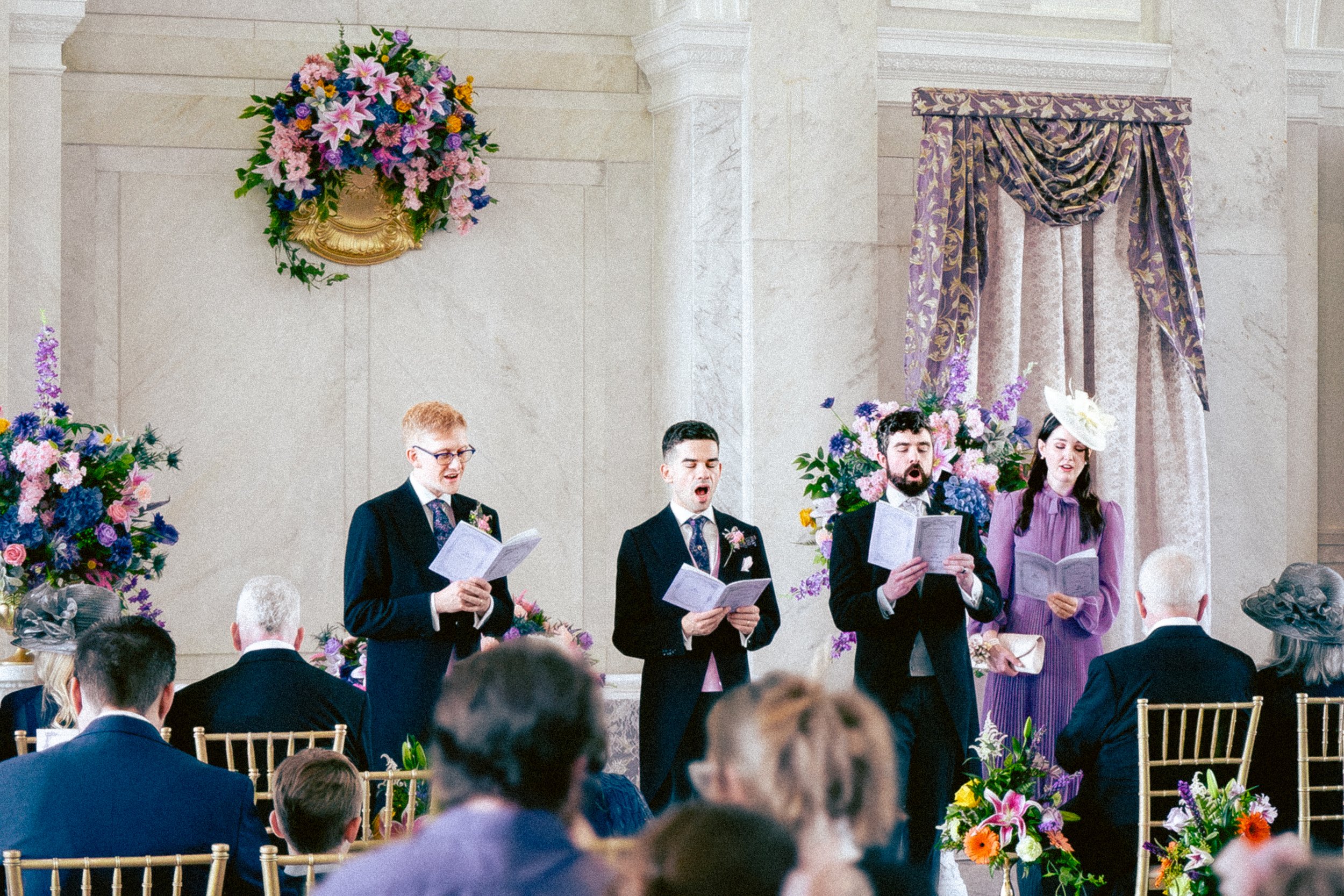 A group of five people singing during a wedding ceremony, standing in front of floral arrangements and a purple-draped backdrop, with wedding guests seated in the foreground.