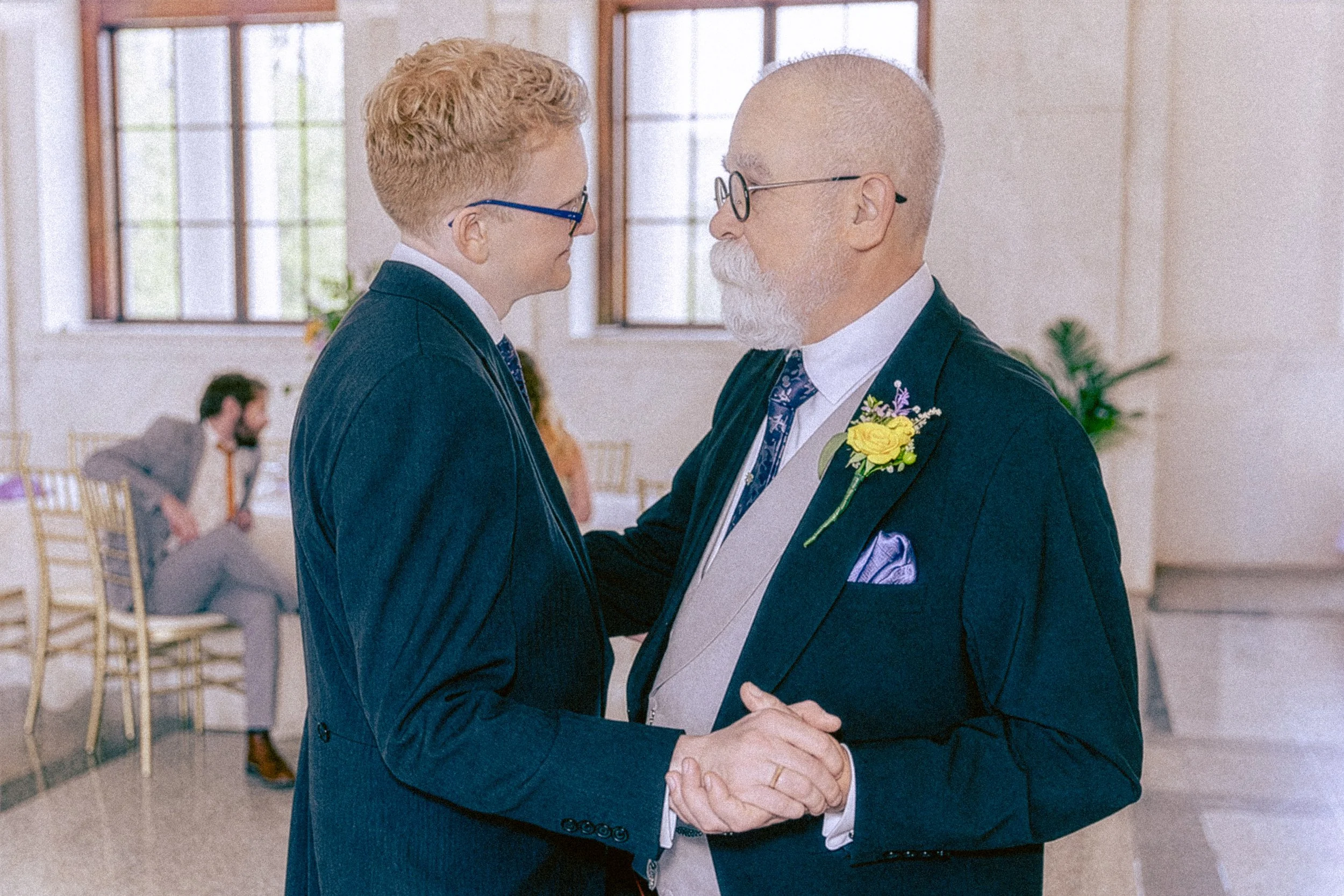 Two men, dressed in suits, are holding hands and gazing into each other's eyes in a bright room with large windows. One man is younger with red hair and glasses, and the other is older with white facial hair and glasses, wearing a boutonniere.
