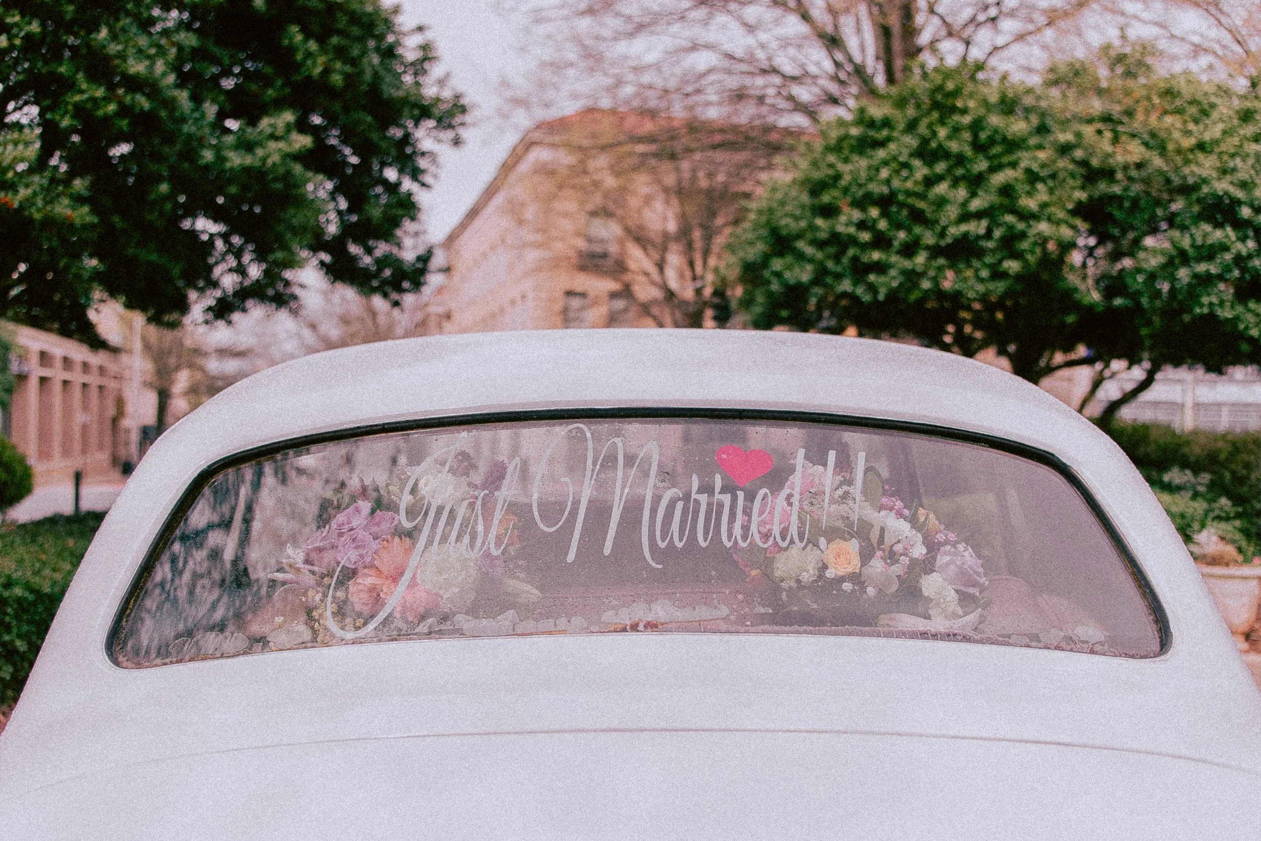 The back of a white vintage car decorated with pink and white flowers and a sign that reads 'Just Married', parked outdoors with trees and buildings in the background.