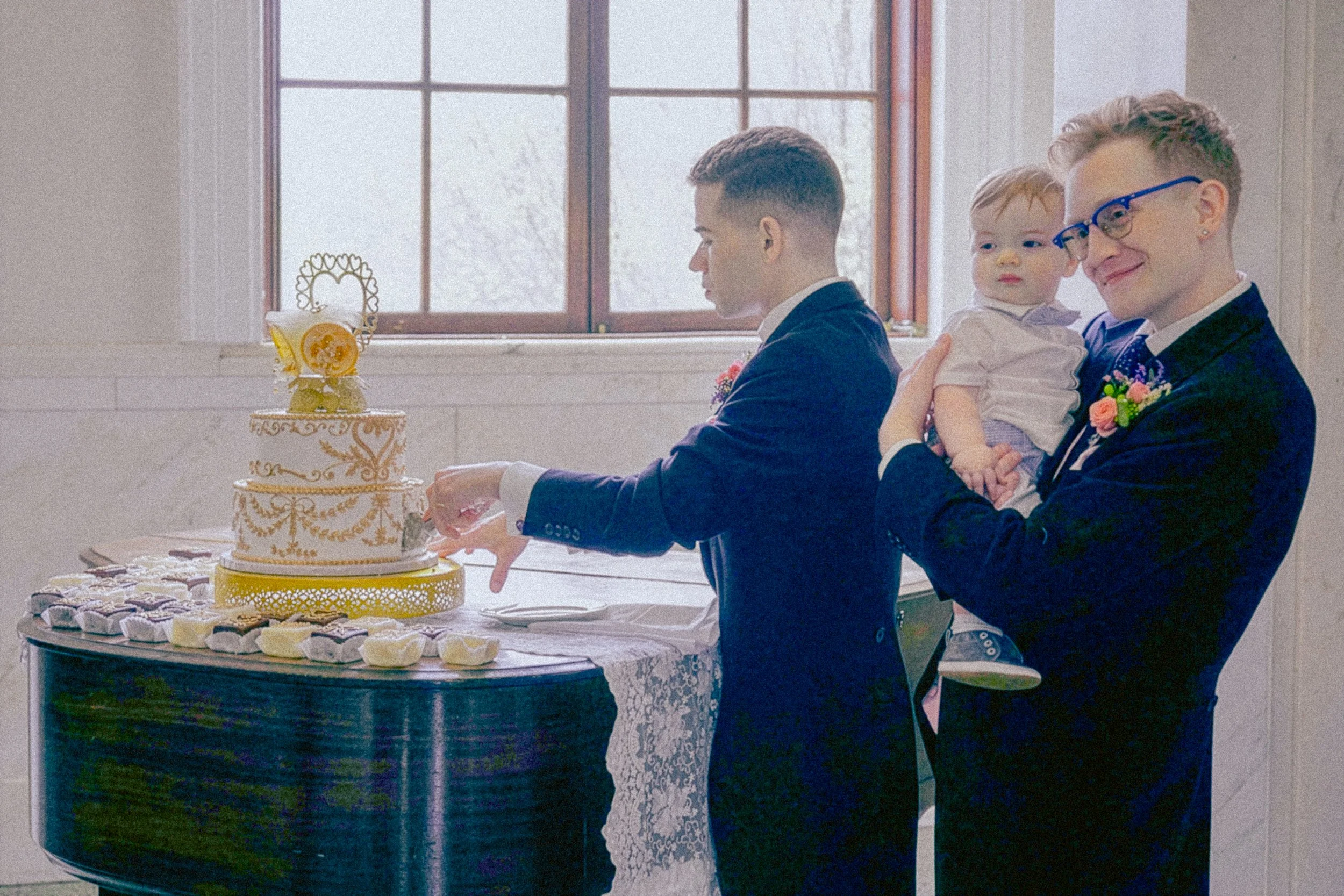 A group of people standing around a table with a two-tiered yellow and white wedding cake on it, decorated with orange slices and floral toppings. The people are in front of a window, and the focus is on a man in a suit slicing the cake while another