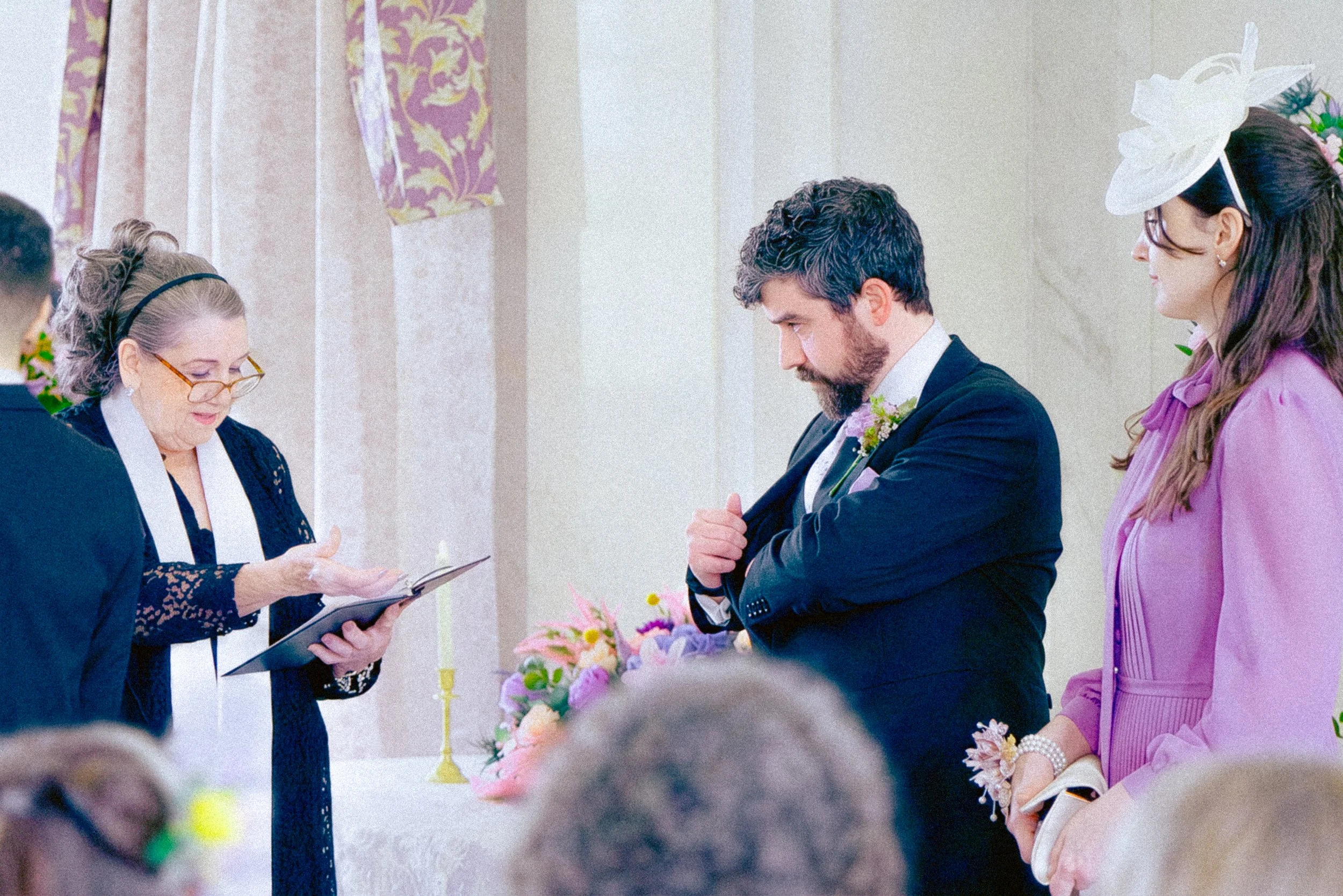 A woman with glasses and a black headband reading from a book during a ceremony, with a man and a woman in formal attire standing nearby, flower arrangements and a candlestick on the table in the background.