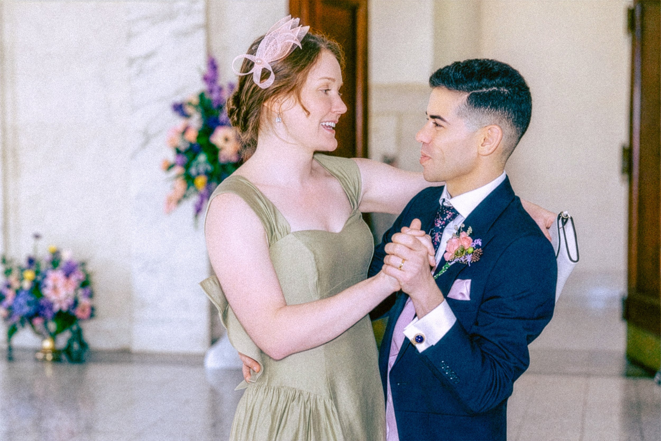 A smiling woman in a light green dress with a pink fascinator is dancing with a man in a dark blue suit at a wedding or formal event. They are holding hands and looking at each other. There are colorful flowers in the background.