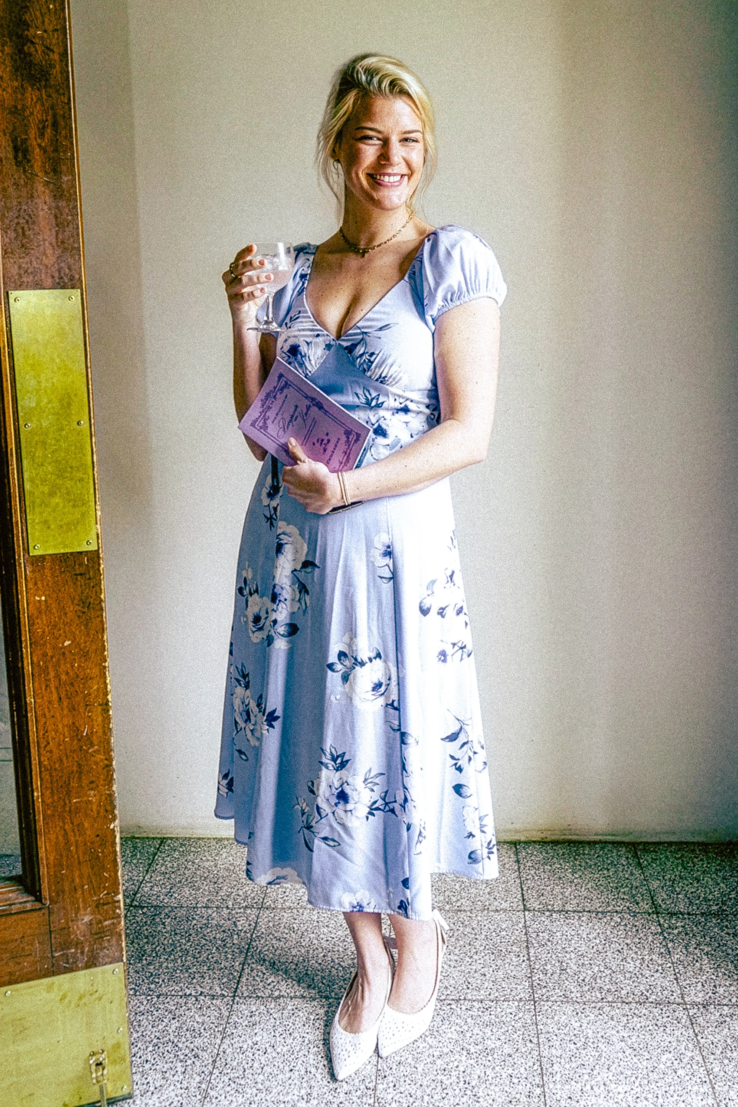 A woman wearing a long, blue floral dress, standing indoors against a beige wall, holding a glass of drink and a pink booklet, and wearing white high heels.