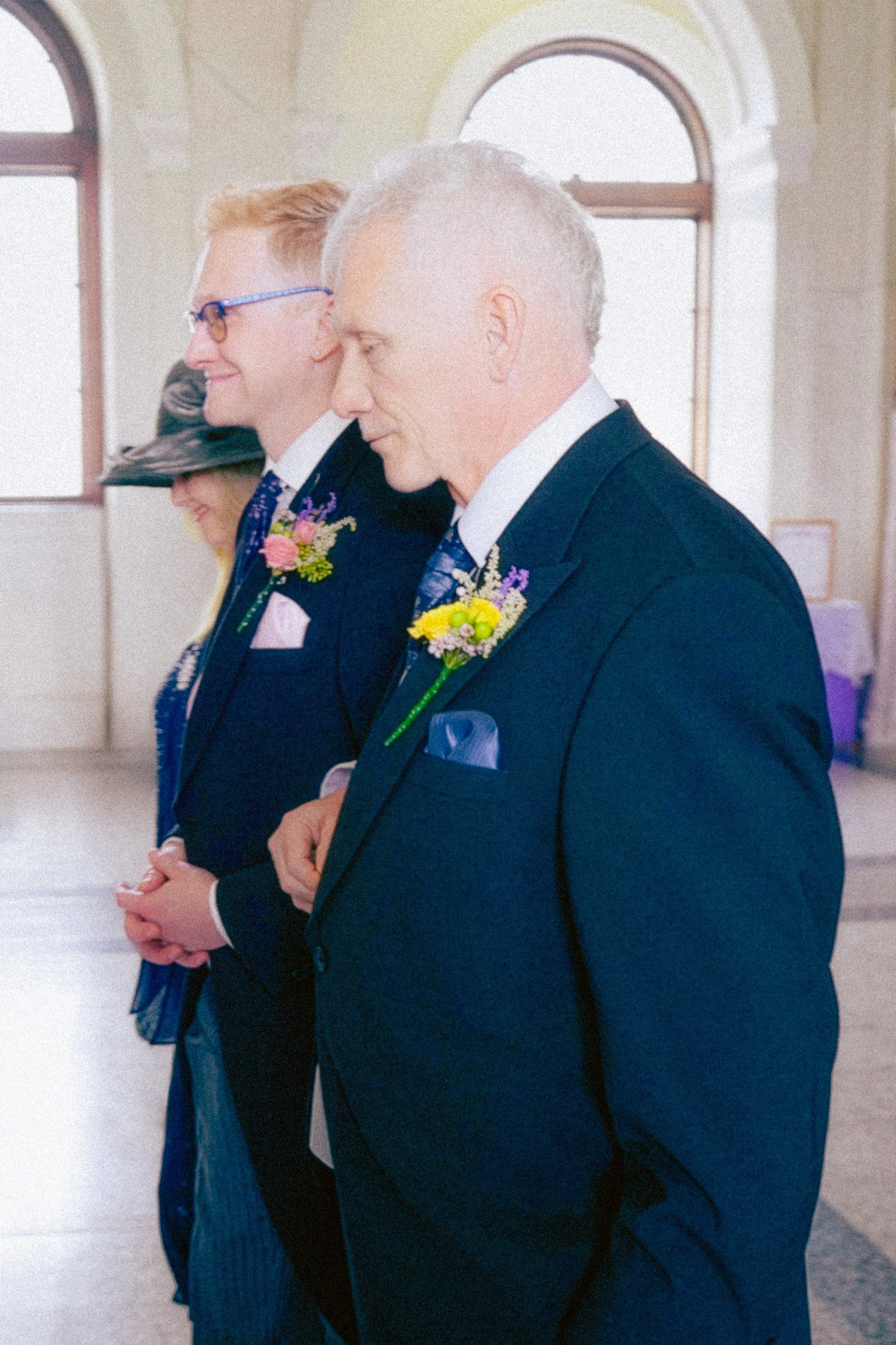 Three individuals in formal attire standing in a line with hands clasped, an older man in the foreground, a young man with glasses and a woman with a hat in the background, all wearing boutonnières, inside a room with large arched windows.