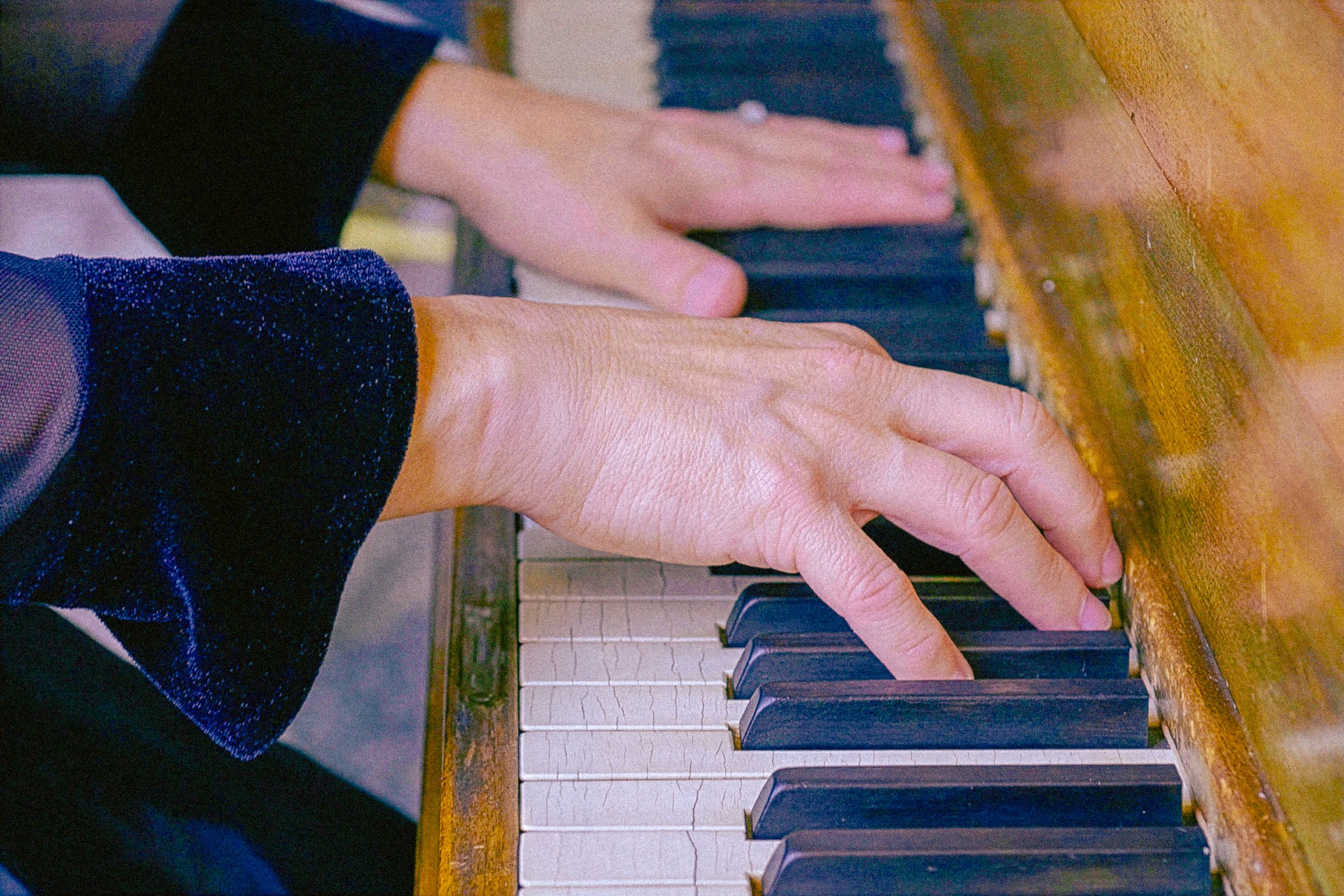A person playing the piano, with their hands on the keys, wearing a dark jacket with a colorful sleeve, and the keys of the piano visible.