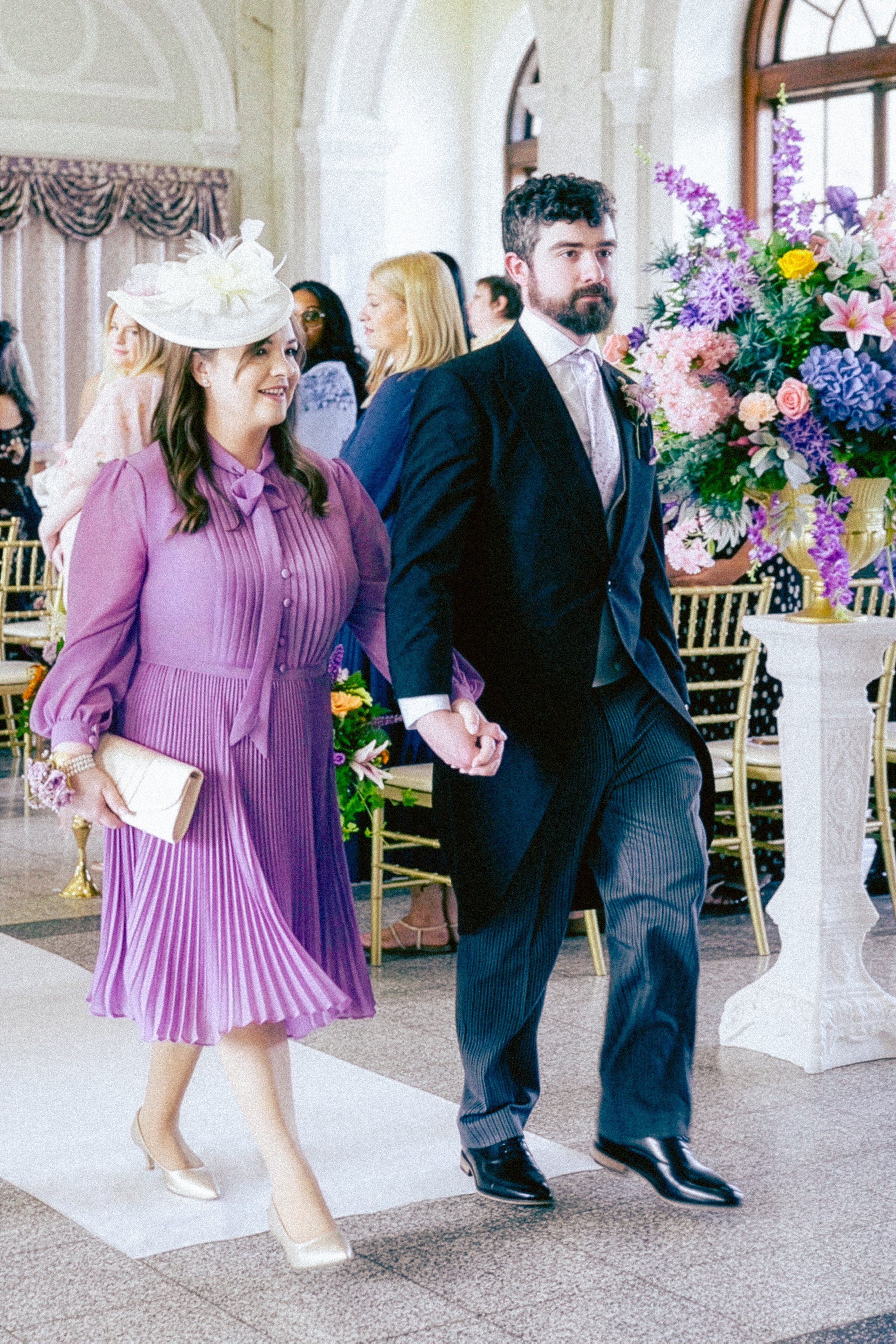 A man and woman are holding hands and walking in a decorated room with floral arrangements, attending a formal event.