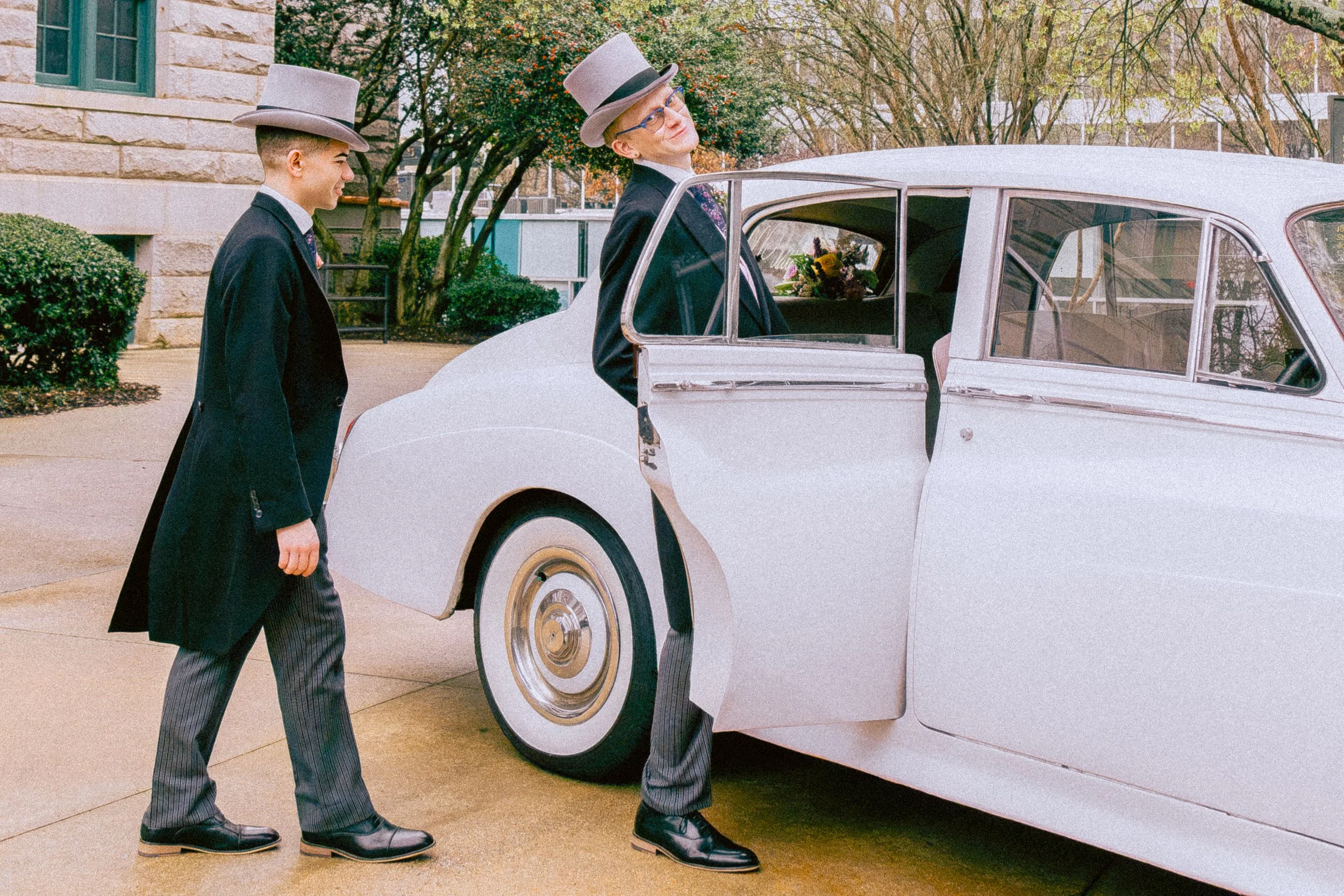Two men dressed in vintage suits and gray top hats, standing next to a white vintage car, with one man smiling and the other looking, in an outdoor setting with trees and buildings in the background.